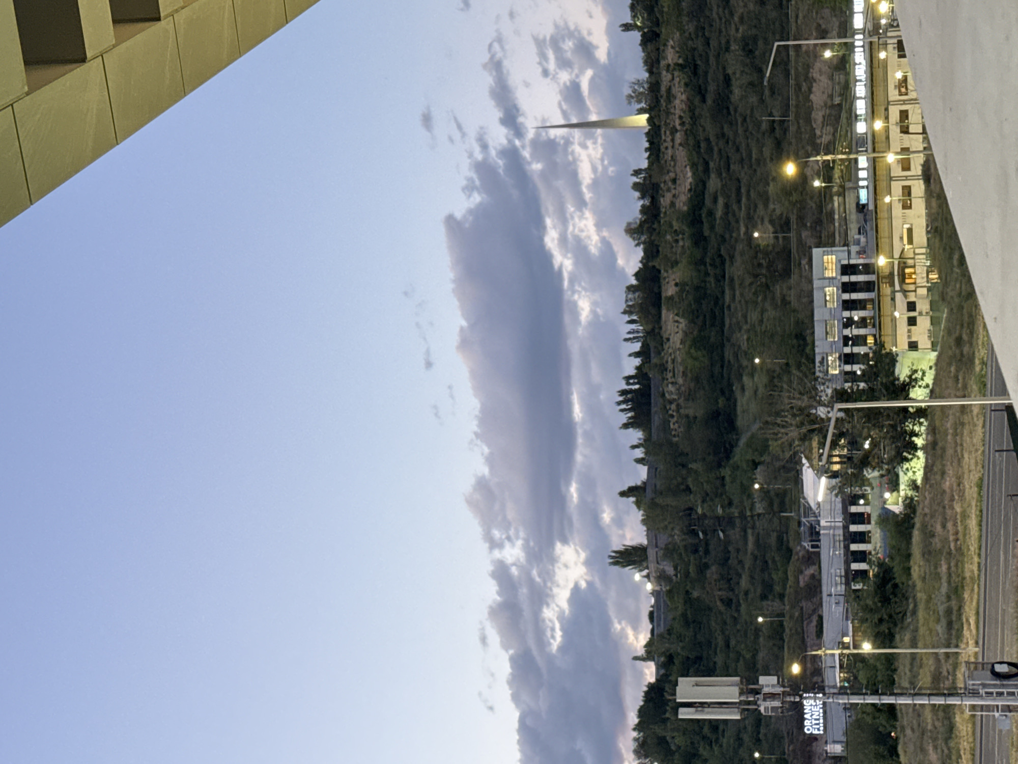 The image, taken at dusk or early evening, presents an urban-adjacent landscape combining natural and architectural elements. The sky, a gradient of light blue, features elongated, partially illuminated clouds. On the right, a prominent green hillside, densely covered with trees, dominates the midground. From this hillside, a tall, slender, light-colored obelisk or spire rises, its apex softly glowing, likely an architectural monument.
Along the base of the hillside, on the lower right, modern buildings with warmly lit windows are visible, interspersed with numerous streetlights that cast a yellow-orange glow across the scene and onto a visible road or pathway. A section of a modern building with a golden-brown facade frames the upper-left corner of the sky. On the far right, a large, lighter-colored structure, possibly a highway overpass or a large building complex, is visible.
In the lower right, a metallic pole structure, appearing to be a cell tower or a large street light pole, holds a vertical sign. The text clearly readable on the sign is "ORANGE FITNESS GYM", with additional, partially obscured text below, possibly "ZANAK..." hinting at a specific location or street name. No people or specific activities are discernible, and the scene conveys a calm, tranquil ambiance as night begins to fall.