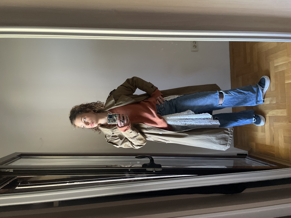 The scene depicts a young adult female captured in a mirror selfie within an indoor setting in Boreti, Montenegro. The female, with light brown, wavy hair and fair complexion, is wearing a long, light brown trench coat over an orange knit sweater, light blue ripped jeans, and black platform sneakers. A white, lace-like fabric is draped from her waist. She holds a smartphone with a camouflage case in her right hand, positioned to capture her reflection, while her gaze is directed slightly to her left, off-camera. Her left hand rests on her hip. The environment consists of a light beige wall visible behind her reflection, a light brown parquet floor arranged in a herringbone pattern, and a European-style double electrical outlet on the lower right wall. To the left, a dark-framed door or closet with a black handle is reflected, indicating its presence opposite the mirror.
