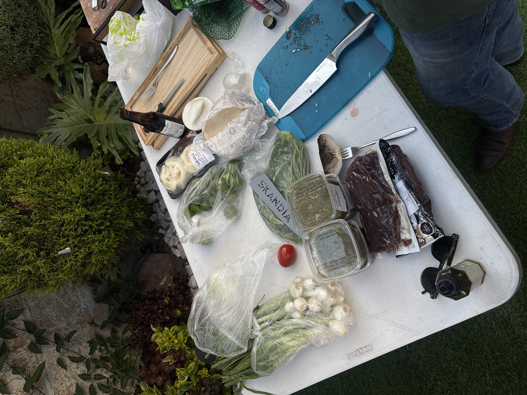 The image captures an outdoor food preparation scene, likely for grilling or a barbecue. The setting appears to be in San Bernardino Tlaxcalancingo, Mexico, as evidenced by the Spanish text on food packaging.
A white folding table is central, laden with various ingredients and utensils. Items include cuts of meat in vacuum-sealed packaging, fresh produce (onions, tomatoes, peppers), a container labeled "SKANDIA," and packaged foods, possibly cheeses. A blue cutting board with a knife, a wooden serving tray with cutlery, and containers holding sauces are also present.
A person wearing jeans and brown shoes is partially visible at the top right, implying they are involved in the food preparation. Other items present are sunglasses and a bottle.
The environment is outdoors, with greenery and ground cover surrounding the table. The presence of rocks and varied plant types indicates a garden or patio setting.