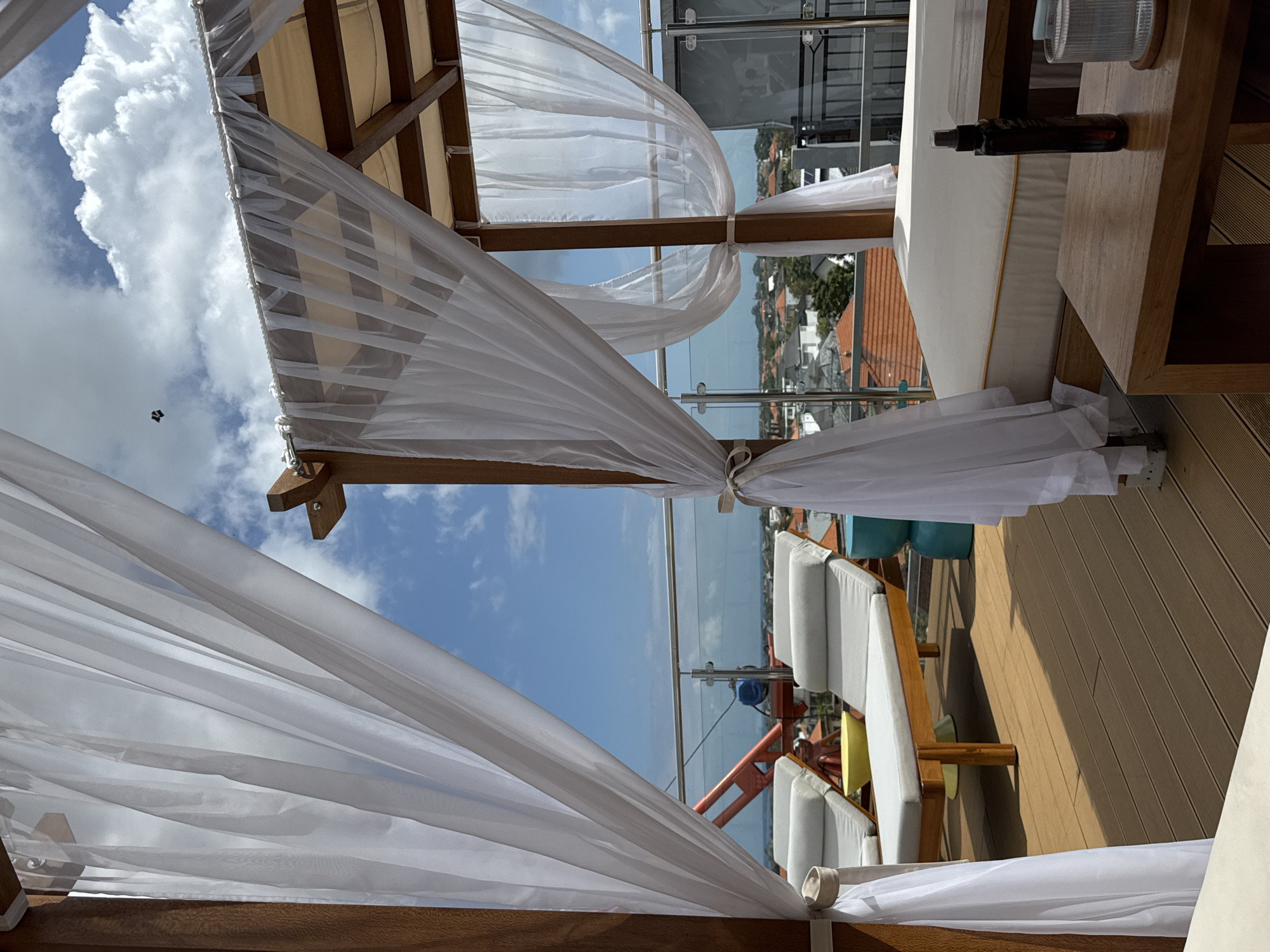 An outdoor patio area, likely a rooftop, in North Kuta, Indonesia, is depicted under a partly cloudy sky. The scene is framed by sheer white curtains draped from a wooden pergola structure.
In the mid-ground, two light gray lounge chairs with wooden frames are arranged on a brown wooden deck. A bright yellow bowl sits on the deck between the chairs. To the right of the chairs, a small turquoise stool is visible. Beyond the chairs, a railing with glass panels overlooks a cityscape of buildings with orange tile roofs.
In the foreground, a low wooden table holds a dark spray bottle and a clear ribbed glass. Adjacent to this table is a large, white mattress with yellow trim, suggesting a daybed or a comfortable seating area.
The overall impression is one of a relaxed, resort-like setting. A small dark object, possibly a kite or a drone, is visible in the upper portion of the sky.