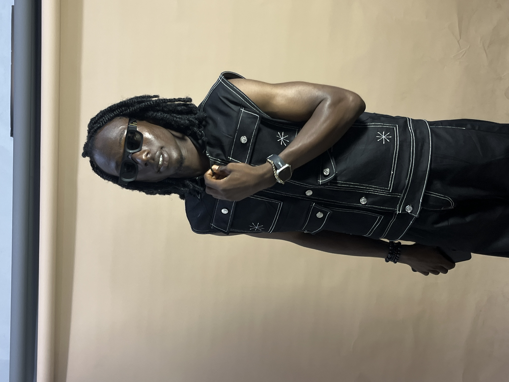 A medium shot shows a man with dark skin and long black dreadlocks wearing dark sunglasses. He is dressed in a black sleeveless vest with white stitching and multiple pockets, a black shirt, and black pants. He is wearing a watch on his left wrist and a bracelet on his right wrist. He is looking at the camera and smiling slightly while holding the front of his vest with his right hand. The background is a plain, light brown. The image is a portrait-style photograph. The time of day, weather, and specific location cues within Gura Top, Nigeria are not discernible from the image. No visible text is present.