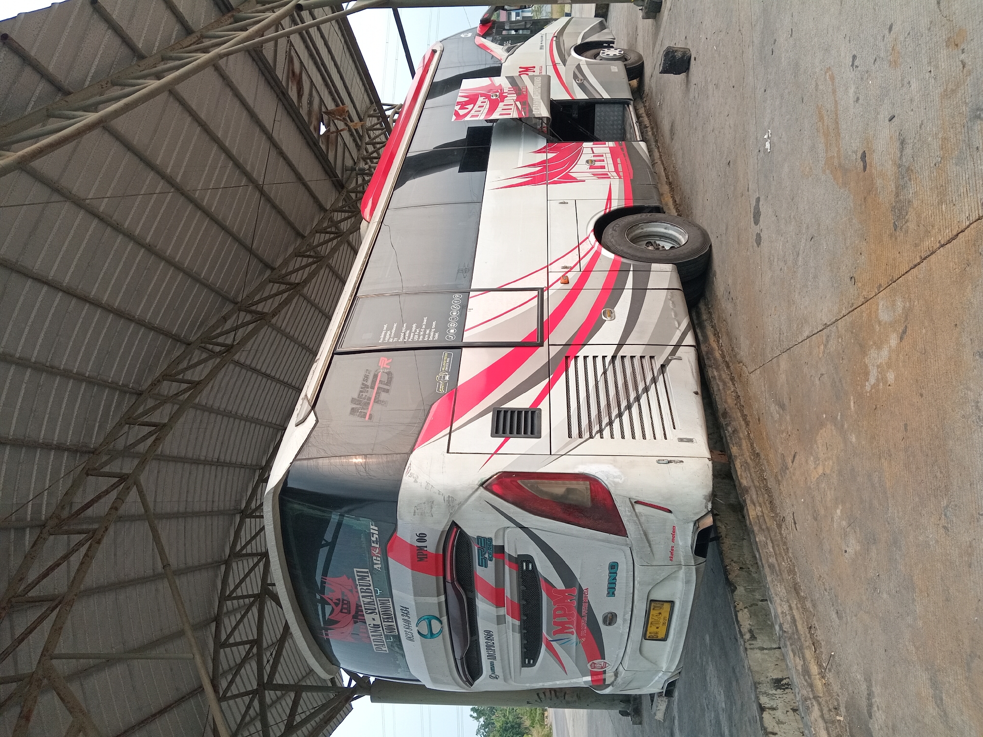 A white and red intercity bus is parked under a metal canopy. The bus has "MPM" branding and "Padang - Sukabumi" written on the back. The front windshield displays "AGRESIP NON EKONOMI". A license plate reading "BA 7036 QU" is visible. The bus's side door is open, revealing storage compartments. The ground is concrete, with some dirt and debris. The environment suggests an outdoor bus station or depot. The image is taken at eye level and appears to be a still photograph.