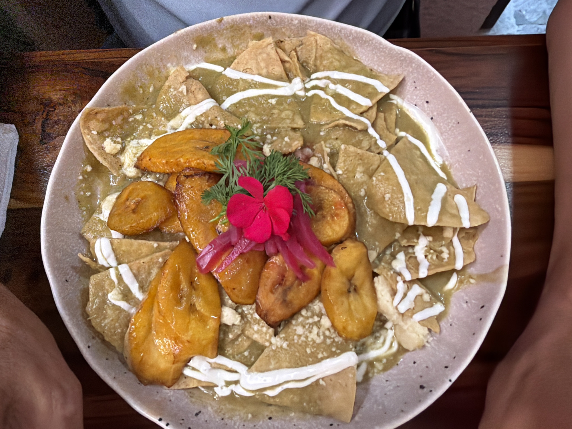 A high-angle shot depicts a plate of chilaquiles with fried plantains, likely taken at a restaurant in an unknown city. The chilaquiles, consisting of tortilla chips coated in green sauce and drizzled with cream, are served on a speckled ceramic plate. At the center of the dish, several golden-brown fried plantain slices are piled, garnished with pickled onions and a small red flower. The plate is placed on a dark, textured wooden table. Two partially visible arms or legs of seated individuals frame the plate on either side.
