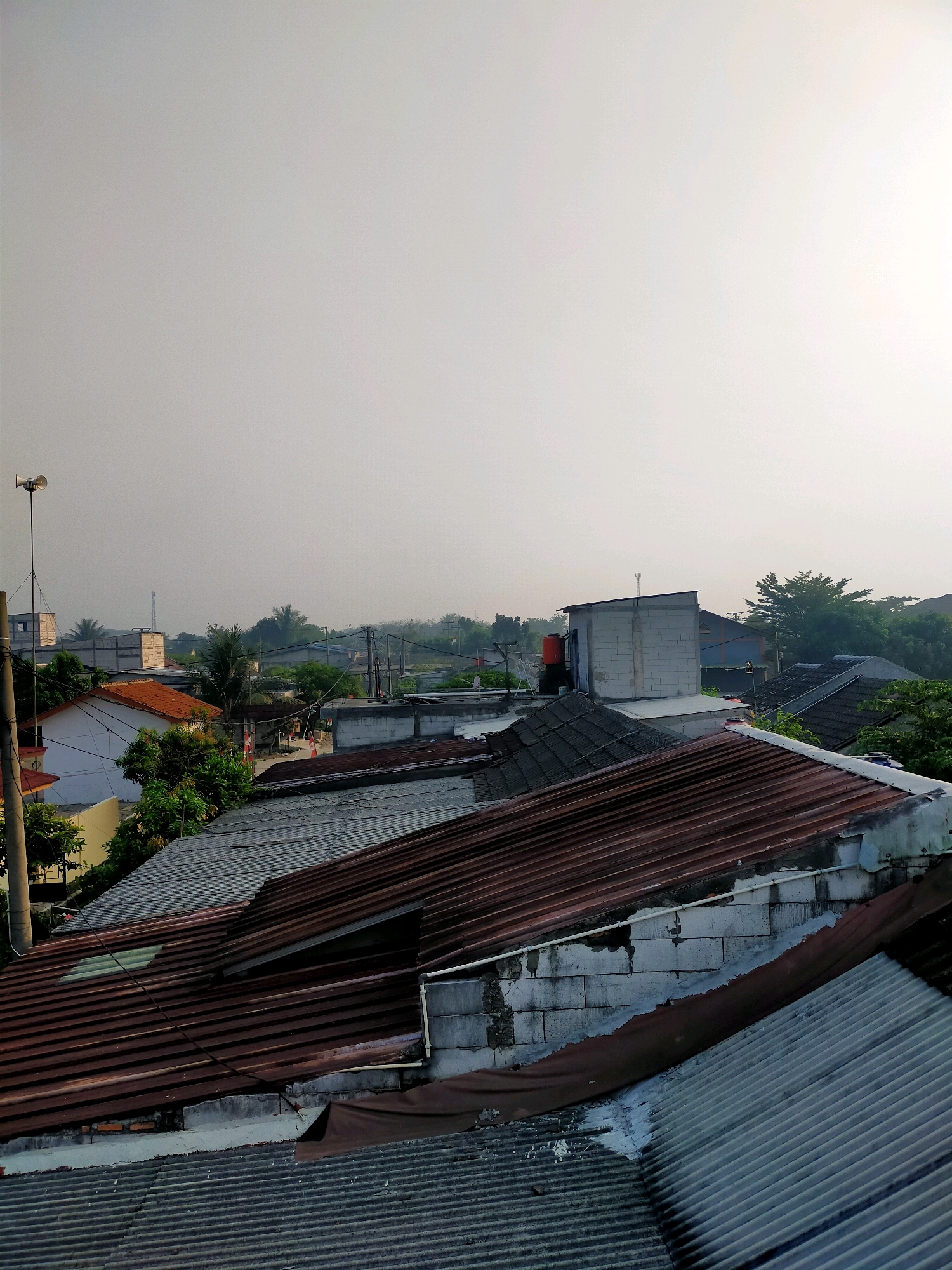 The image depicts a residential area in Sindangmulya, Indonesia, under a hazy or overcast sky. The foreground is dominated by various rooftops, primarily made of corrugated metal, with some showing signs of age and rust. A large, reddish-brown corrugated metal roof occupies a significant portion of the lower and central part of the frame, with a section appearing damaged or detached. To the left and bottom, a grey corrugated metal roof is visible.
In the midground, houses with terracotta-tiled roofs and white walls are interspersed with greenery. Several other buildings, some constructed with concrete blocks, are also present. A tall pole with a speaker and what appears to be a small antenna stands on the left side of the image. Red and white Indonesian flags are visible on some houses.
The background shows a dense arrangement of trees and more distant structures, indicating a densely populated urban or suburban environment. The overall impression is of a typical neighborhood scene in Indonesia, with a focus on the architectural elements of residential buildings and their roofing materials, set against a muted, hazy sky. There are no people visible in the image.