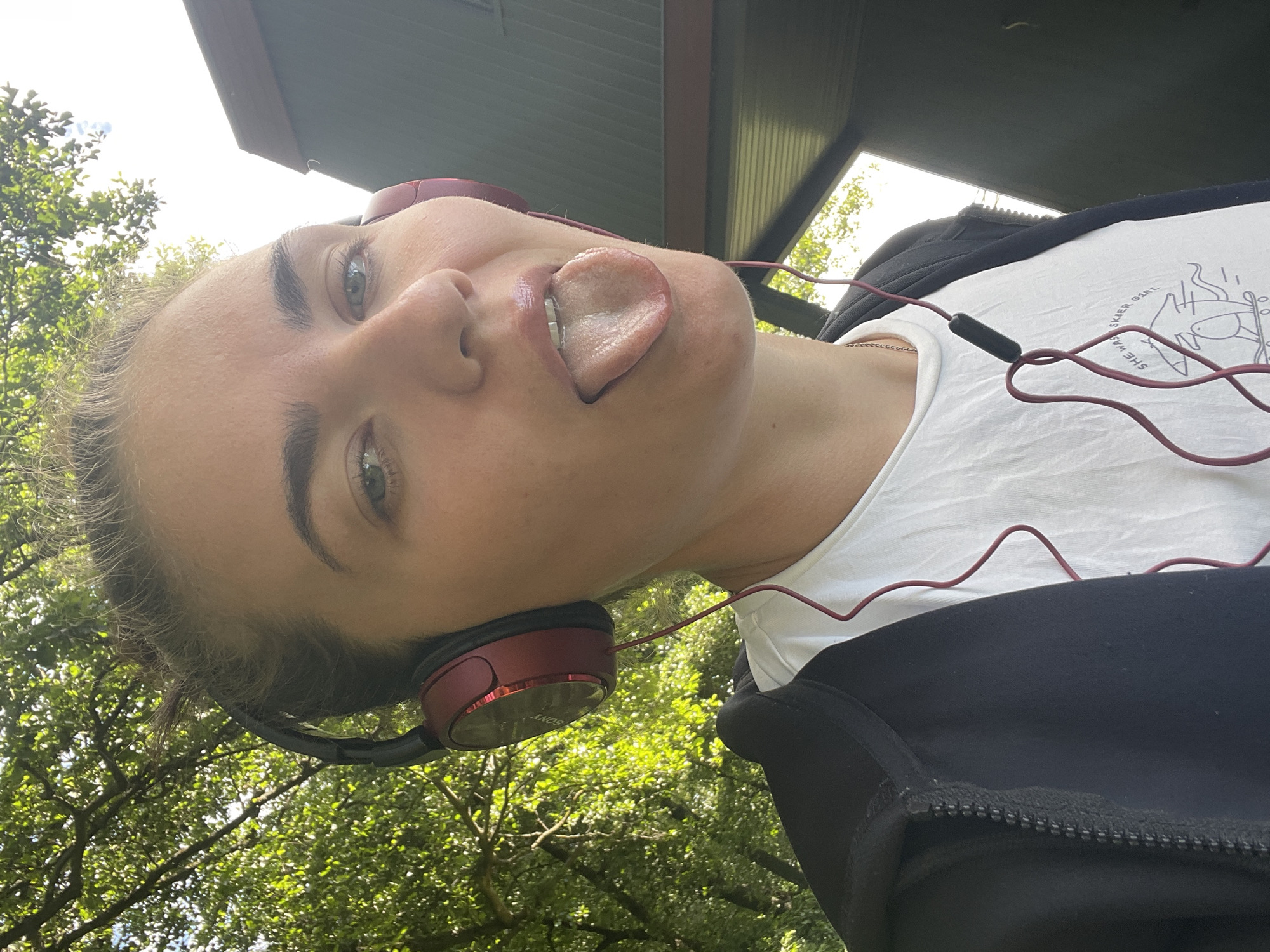 A young woman wearing headphones and a black zip-up hoodie over a white t-shirt sticks her tongue out at the camera. Her hair is pulled back in a bun. The background shows green trees and a structure with a dark roof. The image appears to be taken outdoors during the day. The location is not specified in the image.