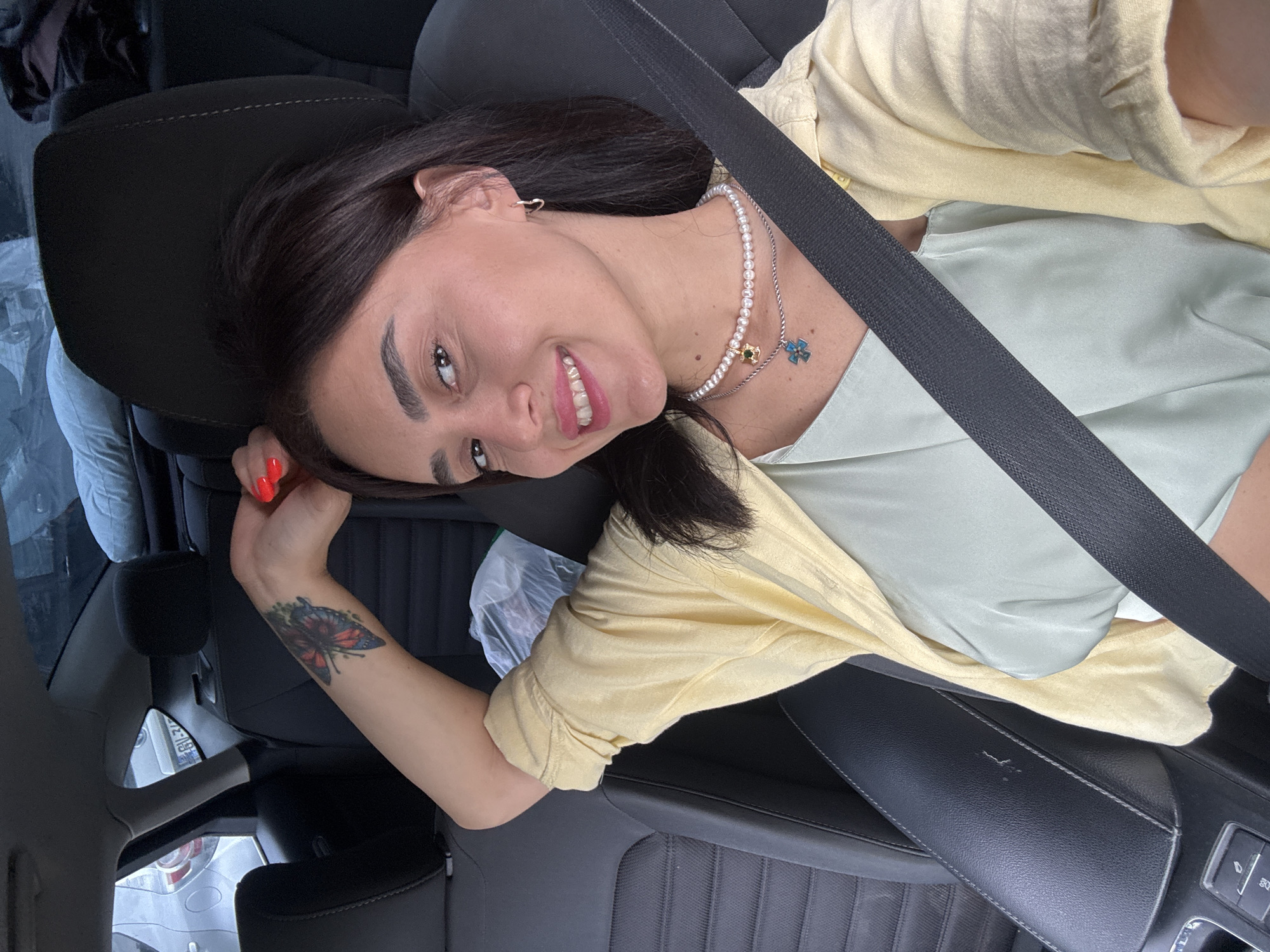 A woman with dark hair and bright red nail polish is captured in a selfie taken from inside a car. She is smiling and wearing a light yellow open-collar shirt over a pale green sleeveless top. A pearl necklace with a green charm and a silver chain with a blue charm are visible. She has a butterfly tattoo on her left arm, which is resting behind her head. A seatbelt crosses her chest. The background shows the interior of the car, including the headrests and seats. Outside the car windows, other vehicles and parts of what appears to be a parking lot are visible. The scene is consistent with a common urban setting, and the image does not provide any specific visual cues to pinpoint the location as Tbilisi, Georgia.