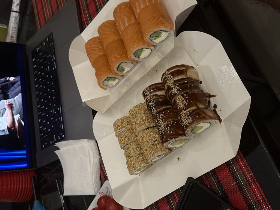 This image depicts an indoor scene in Budva, Montenegro, featuring food and digital media. No individuals are directly visible within the physical scene.
In the foreground, two white cardboard takeout containers hold a variety of sushi rolls. The container on the right features six rolls uniformly wrapped in salmon, with their cross-sections revealing a creamy white filling and a green interior, consistent with cream cheese and avocado or cucumber. The container on the left contains twelve rolls: six are generously coated in sesame seeds, and the other six are drizzled with a dark, glossy sauce, likely eel sauce, and topped with sesame seeds. These rolls also display creamy white and green interiors. A small, black rectangular dish holding a dark liquid, consistent with soy sauce, is positioned to the lower left of the sushi containers.
In the mid-ground, a dark grey laptop with an illuminated keyboard is open, its screen displaying two adult males in a conversation or interview setting; one is wearing a light-colored shirt, and the other a dark suit. To the immediate left of the laptop, a stack of clean white paper napkins rests on the surface, adjacent to a small white dish holding approximately four ripe red cherry tomatoes.
All depicted objects are arranged on a red and green plaid textile surface, consistent with a tablecloth or placemat, which overlies a darker, possibly black, underlying surface. The lighting suggests an indoor, possibly evening, environment.