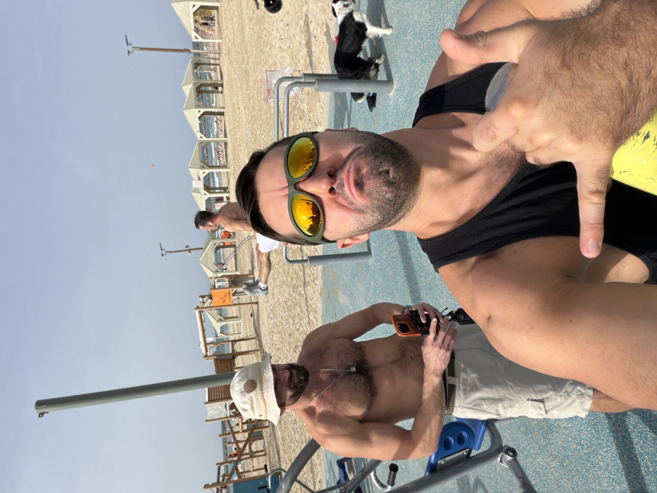 The image depicts an outdoor scene at a beach in Tel Aviv, Israel, specifically in an area featuring public exercise equipment adjacent to the sand. In the foreground, a male subject is taking a selfie. He has short dark hair, a beard, and wears dark green-framed sunglasses with orange mirrored lenses and a black tank top. His mouth is pouted, and his right hand is partially visible, making a gesture with his thumb and forefinger extended, while holding a clear bottle.
To the left and slightly behind the foreground subject, a second shirtless male stands with a beard and a light-colored bucket hat. He wears light-colored cargo shorts and a silver chain with a pendant. He holds an orange smartphone in his hands and is looking towards the selfie-taker.
Further back on the right side of the frame, a third shirtless male is performing an exercise on a horizontal bar apparatus, with his legs extended horizontally. He wears white shorts and grey sneakers. A black and white dog with distinct markings walks on the sandy beach in the mid-ground, near the exercise area.
The background features a wide sandy beach with numerous light-colored, square beach canopies extending towards the calm, blue Mediterranean Sea under a clear, light blue sky. Several vertical poles are also visible across the beach landscape. The exercise equipment is situated on a blue rubberized mat.