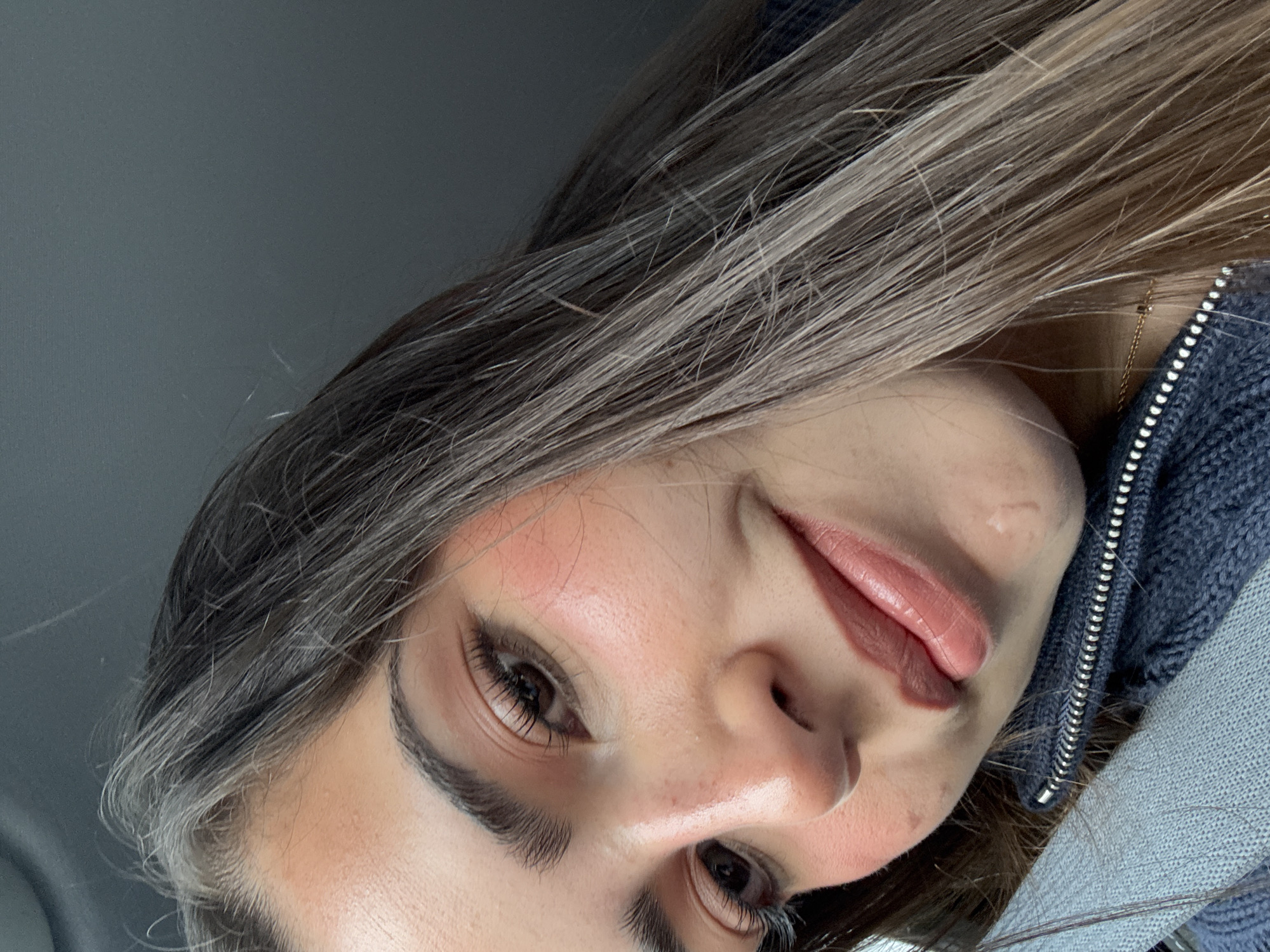 A close-up, tilted shot of a woman with long, light brown hair and fair skin. She has dark eyes with long lashes. She is wearing a blue knit sweater or jacket. A gold necklace is visible at her neck. The background is a light gray color, possibly a ceiling or interior wall. The location is not determinable from the image.