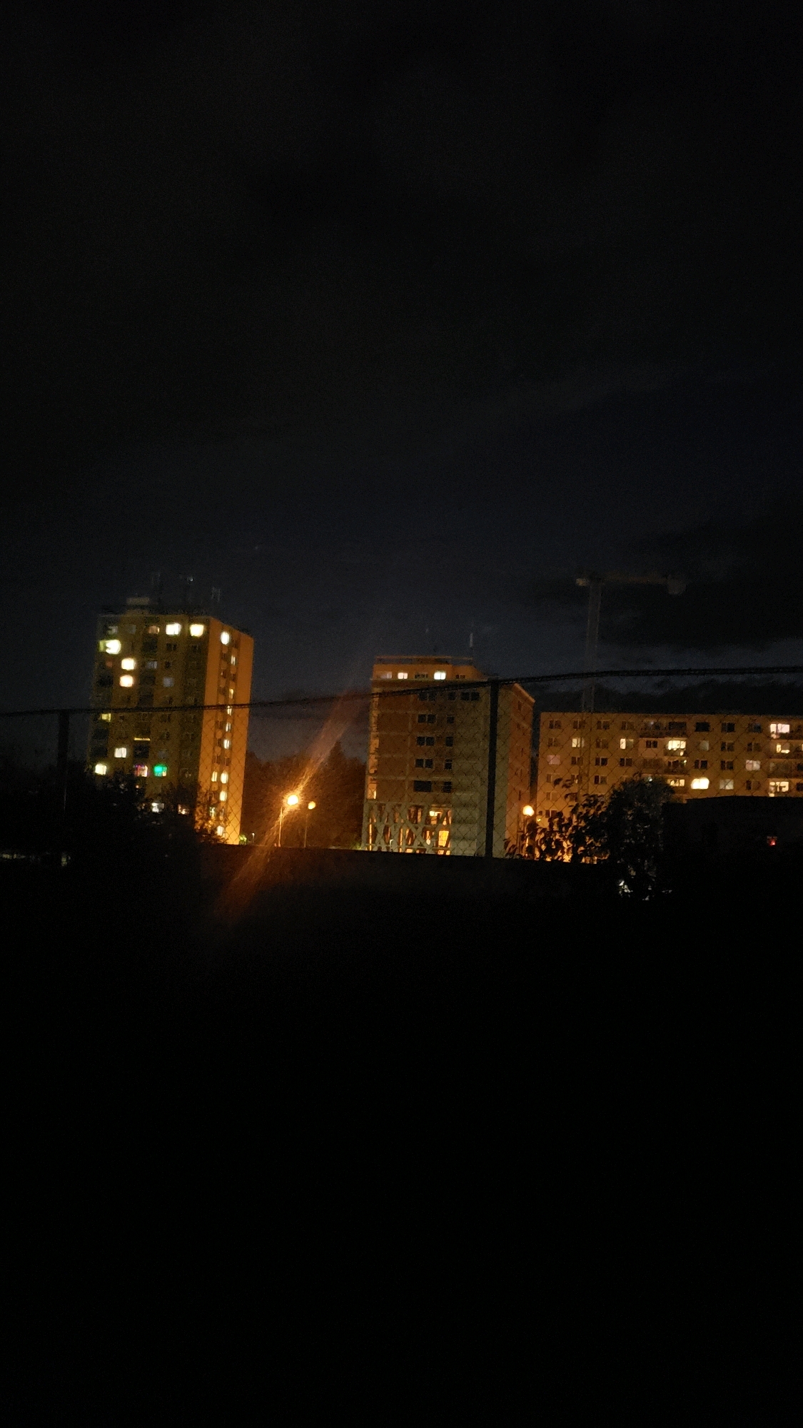 The image captures a night scene in the city of Prešov, Slovakia, likely in a residential area with high-rise buildings. Two apartment buildings are visible, illuminated by numerous lit windows, suggesting occupancy. The buildings appear to be multi-story structures, possibly Soviet-era panelák-style apartment blocks, common in Eastern European cities. In the background, a construction crane is faintly visible against the dark sky, indicating ongoing development or renovation in the vicinity. The foreground is dominated by darkness, with a blurred fence or barrier creating a sense of distance or separation from the buildings. A street lamp with a warm, orange glow illuminates the area between the buildings. The overall atmosphere is nocturnal and urban, with the lights from the apartments providing the primary source of illumination.