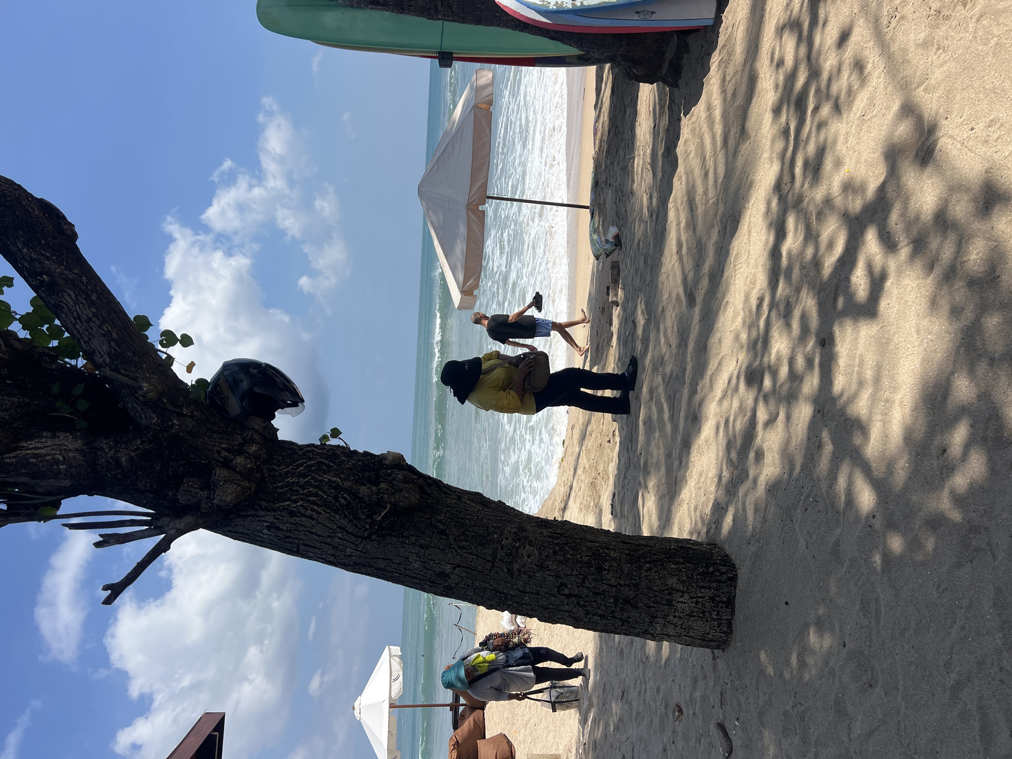 A beach scene in Kuta, Indonesia features a sandy shore with several people and objects. In the foreground, a large tree trunk is prominent, with shadows cast by its leaves on the sand. To the left, a person wearing a blue hat and grey shirt is walking with a bag, past a cluster of brown beanbag chairs and a white umbrella. Further back, two people are walking towards the ocean. One, wearing a yellow shirt and black hat, is carrying a bag. The other, a man in blue shorts and a dark t-shirt, is holding flip-flops. A light-colored umbrella stands near the water's edge. The ocean has moderate waves, and the sky is blue with scattered white clouds. On the right side of the image, two surfboards, one green and one with blue and white patterns, are leaning against a dark structure, likely a tree. A black helmet is attached to the tree trunk in the foreground.