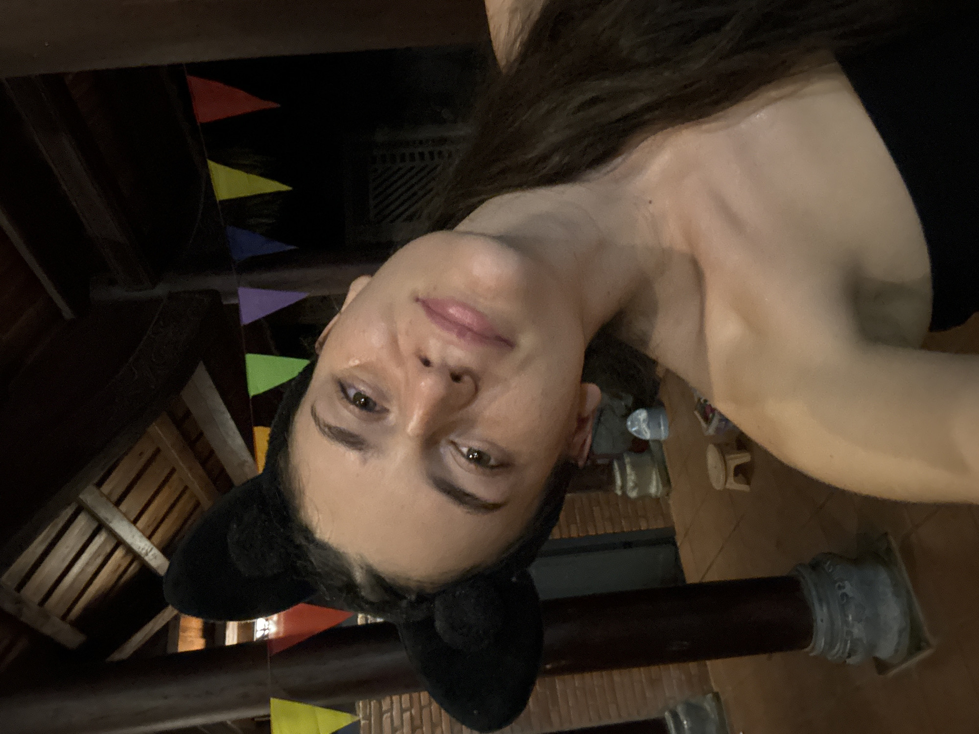 The image shows a woman with dark hair and black cat-ear headbands, positioned at an angle, seemingly indoors in Mũi Né, Vietnam. Triangular colored pennants are strung overhead. The location appears to be an open-air structure with wooden beams, possibly a patio or terrace. Concrete pillars are also visible, as is a floor covering. In the background, there are glimpses of a table and chairs. The subject is wearing a black top, and her skin appears slightly damp.