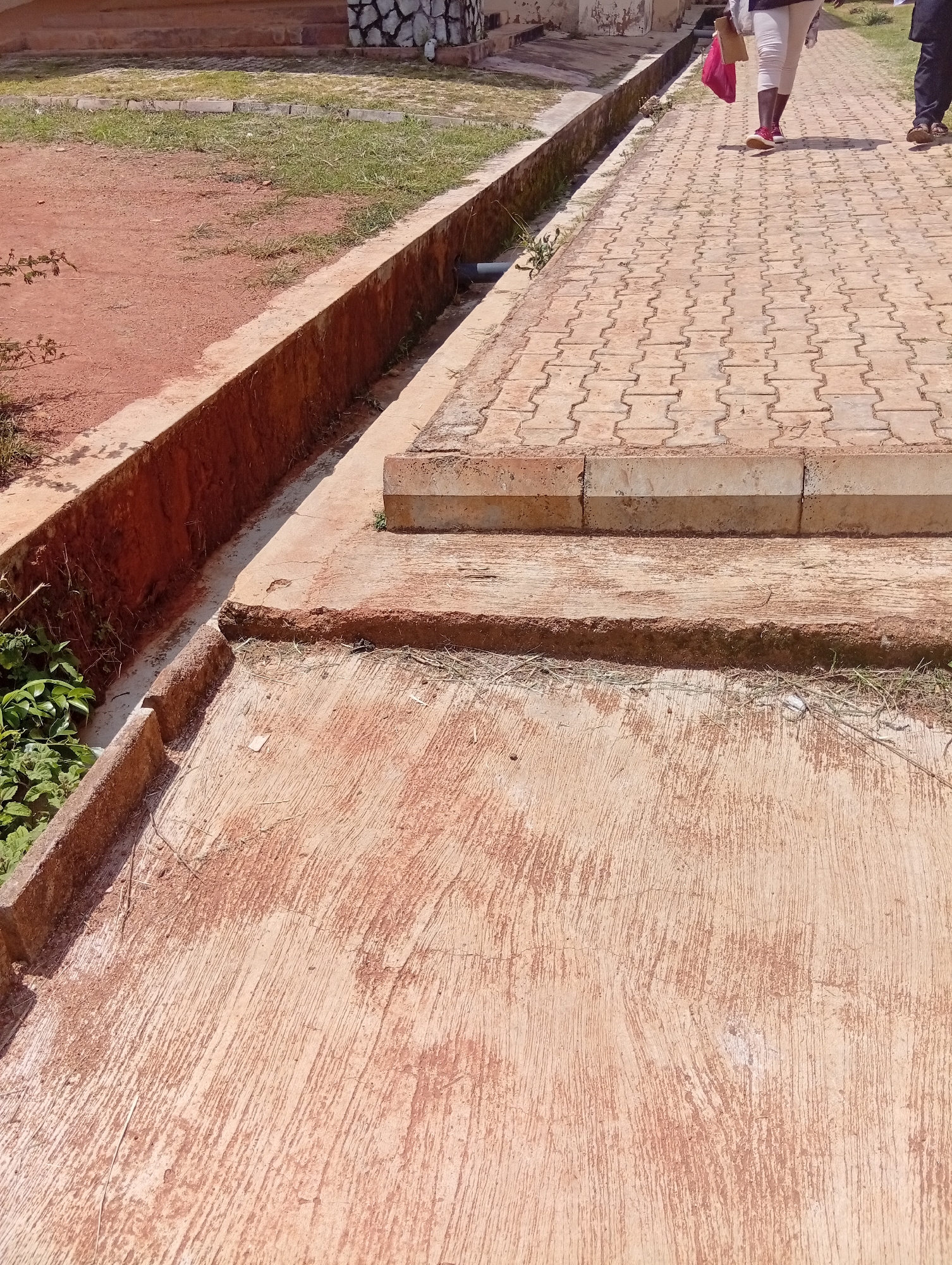 The image captures an outdoor scene in Gwong, Nigeria, featuring two individuals walking on a paved walkway. The walkway is constructed from interlocking brick pavers in a tan color, arranged in a repeating pattern. To the left of the walkway, there is a concrete drainage channel with reddish-brown earth and some greenery growing along its edges. Beyond the drainage channel, a dirt area with sparse grass is visible, leading towards a building with stepped brick foundations. The building's facade appears to be decorated with light-colored stones.
The two people are seen from the waist down, walking away from the viewer. The person in front is wearing white trousers and red sneakers, carrying a red bag. The person behind them is wearing dark trousers and sandals. The lighting suggests it is daytime, with shadows present, indicating a sunny or partly cloudy day. The overall impression is of a functional outdoor space, possibly a public area or pathway on a campus. There is no visible text in the image.