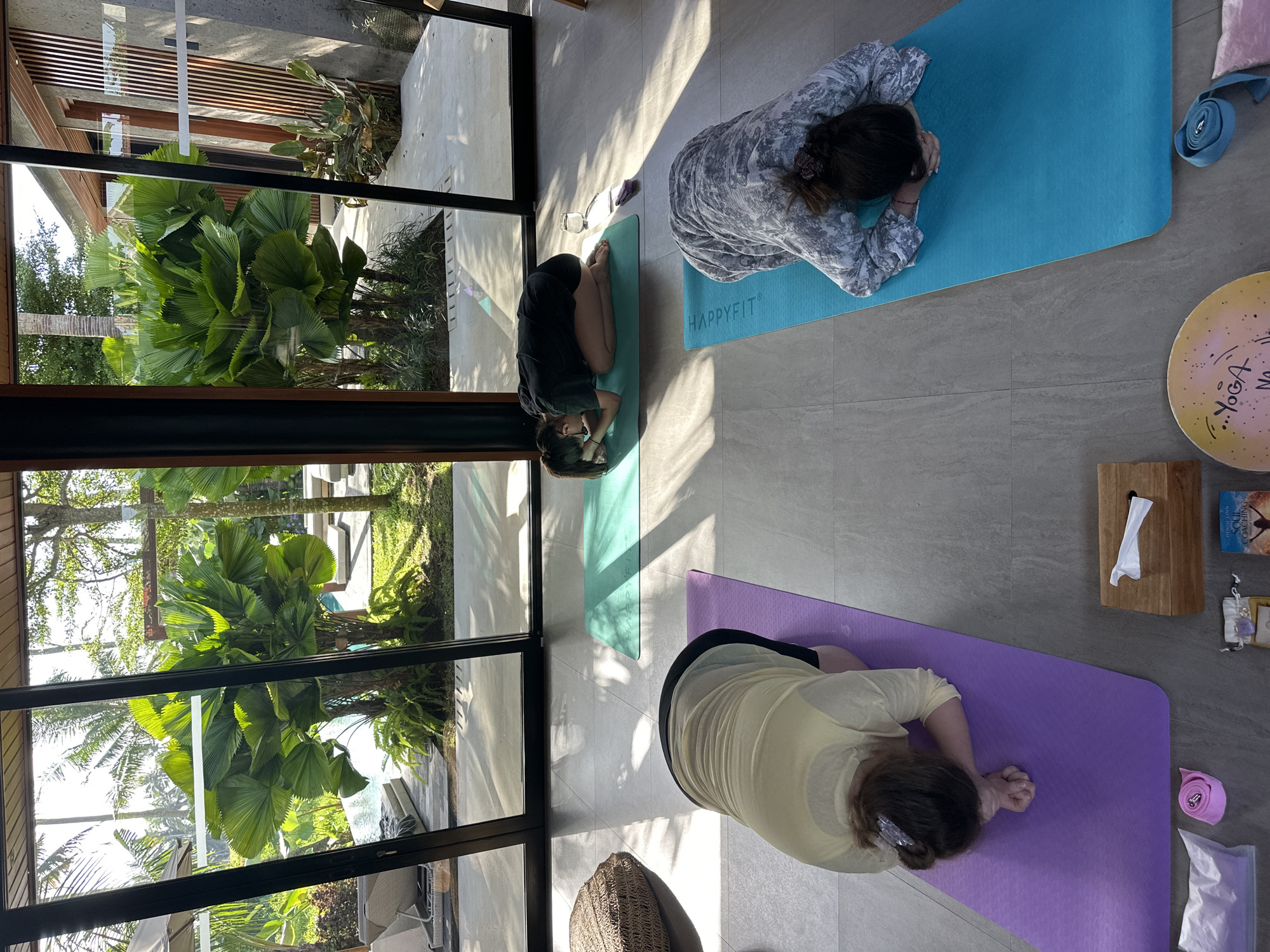 Three people are doing yoga in an indoor space in Sukawati, Indonesia. The space has large floor-to-ceiling windows that provide views of lush tropical foliage outside. The yoga practitioners are positioned on individual mats: one on a purple mat, another on a teal mat with "HAPPYFIT" printed on it, and the third on a green mat. All three individuals are in a prone position with their foreheads resting on the mat. Nearby, there is a pink yoga strap, a wooden tissue box, and a round yoga board with "YOGA" written on it. Outside, the landscape is filled with dense tropical plants and trees, reflecting the region's typical vegetation. The indoor space is illuminated by natural light filtering through the windows. The floor is tiled in a neutral tone. A small woven basket sits near the purple mat. A yoga block and a book titled "Soul Coaching" are also visible near the purple mat.
