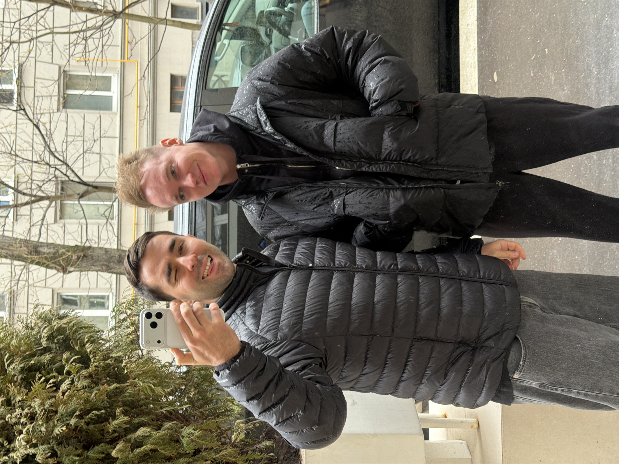 Two men are posing for a selfie outdoors. One man is holding a smartphone and smiling. The second man is standing behind him, also smiling. Both men are wearing black puffer jackets. The background includes a car, a building with multiple windows, bare tree branches, and a green bush. Based on the architecture and vegetation, the location appears to be in an urban area, possibly Moscow, Russia.