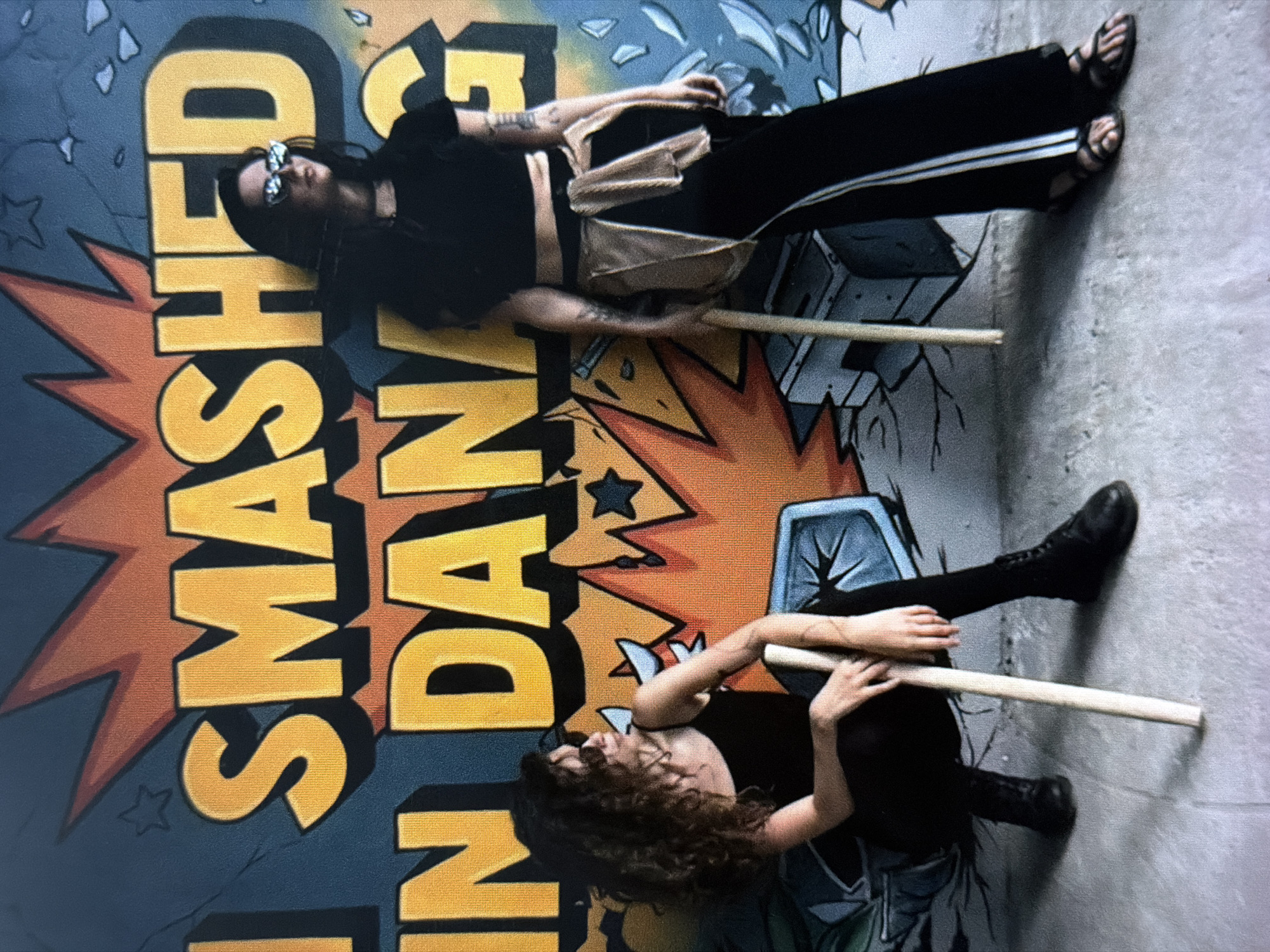 Two people are lying down in front of a mural that reads "UNLEASHED SMASHED." The person on top is wearing sunglasses, a black shirt, black pants with a white stripe, and sandals. They also have a light-colored bag slung across their body. The person on the bottom is wearing a black shirt, black pants, and black boots. Both people are holding a wooden stick. The location is not visible. The location is most likely within the city of Da Nang, Vietnam.