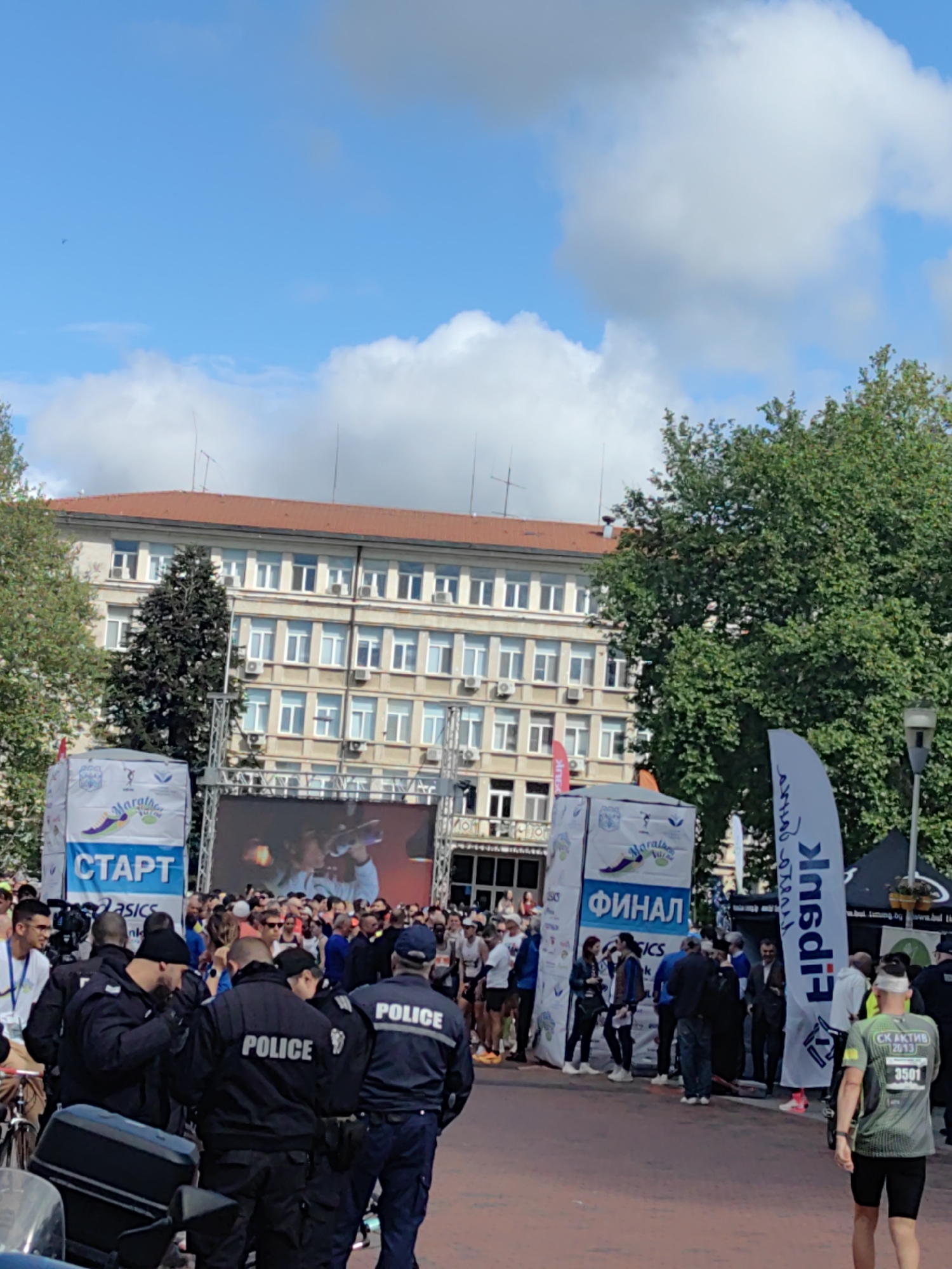 The image captures a scene in what appears to be the city of Varna, Bulgaria, during a marathon event. In the foreground, there is a group of police officers, with the word "POLICE" clearly displayed on their jackets. The crowd of people in the background appears to be a mix of participants and spectators. The banners with "СТАРТ" (Start) and "ФИНАЛ" (Finish) in Cyrillic indicate the beginning and end points of the marathon race. A large screen is set up behind the starting line, possibly showing the race or related content. The buildings in the background suggest an urban setting with a mix of architectural styles. The weather seems pleasant with a blue sky and some clouds. Runners can be seen near the starting line, ready for the race, while others are near the finish line. A man wearing a green and black running outfit with the number 3501 is visible in the lower right corner.
