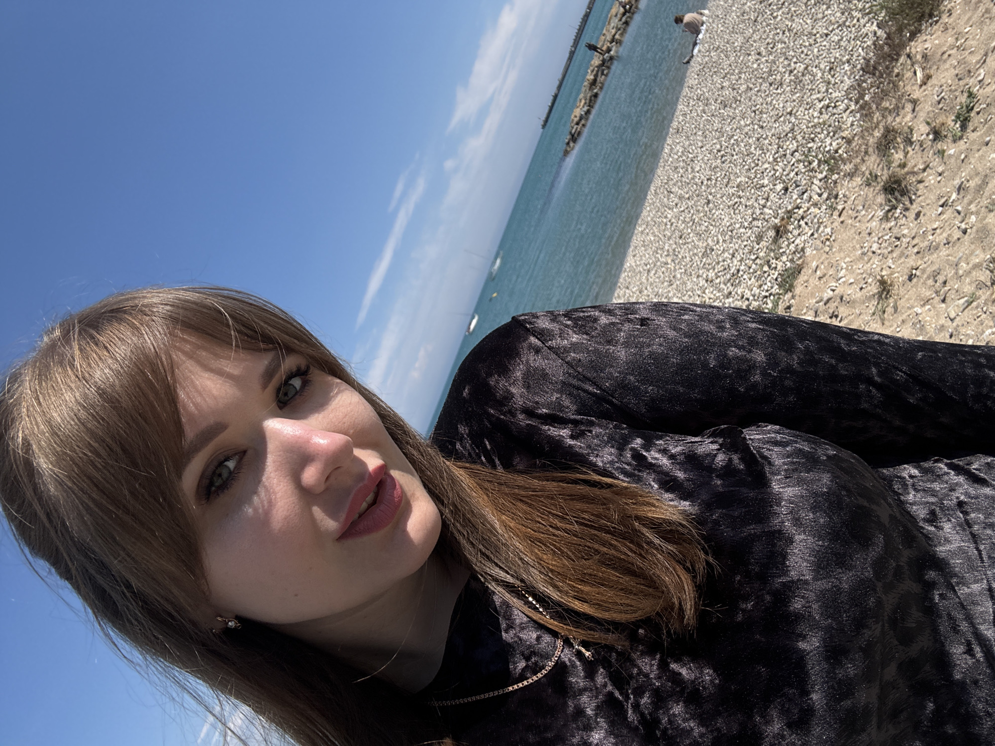 A woman is taking a selfie at a slight angle. The location is a beach in Yevpatoriya, Ukraine, characterized by small rocks near the water's edge and a pier or breakwater in the distance. The sky is blue with some clouds. The woman has light brown hair, fair skin, and is wearing a dark-colored velvet-like shirt.
