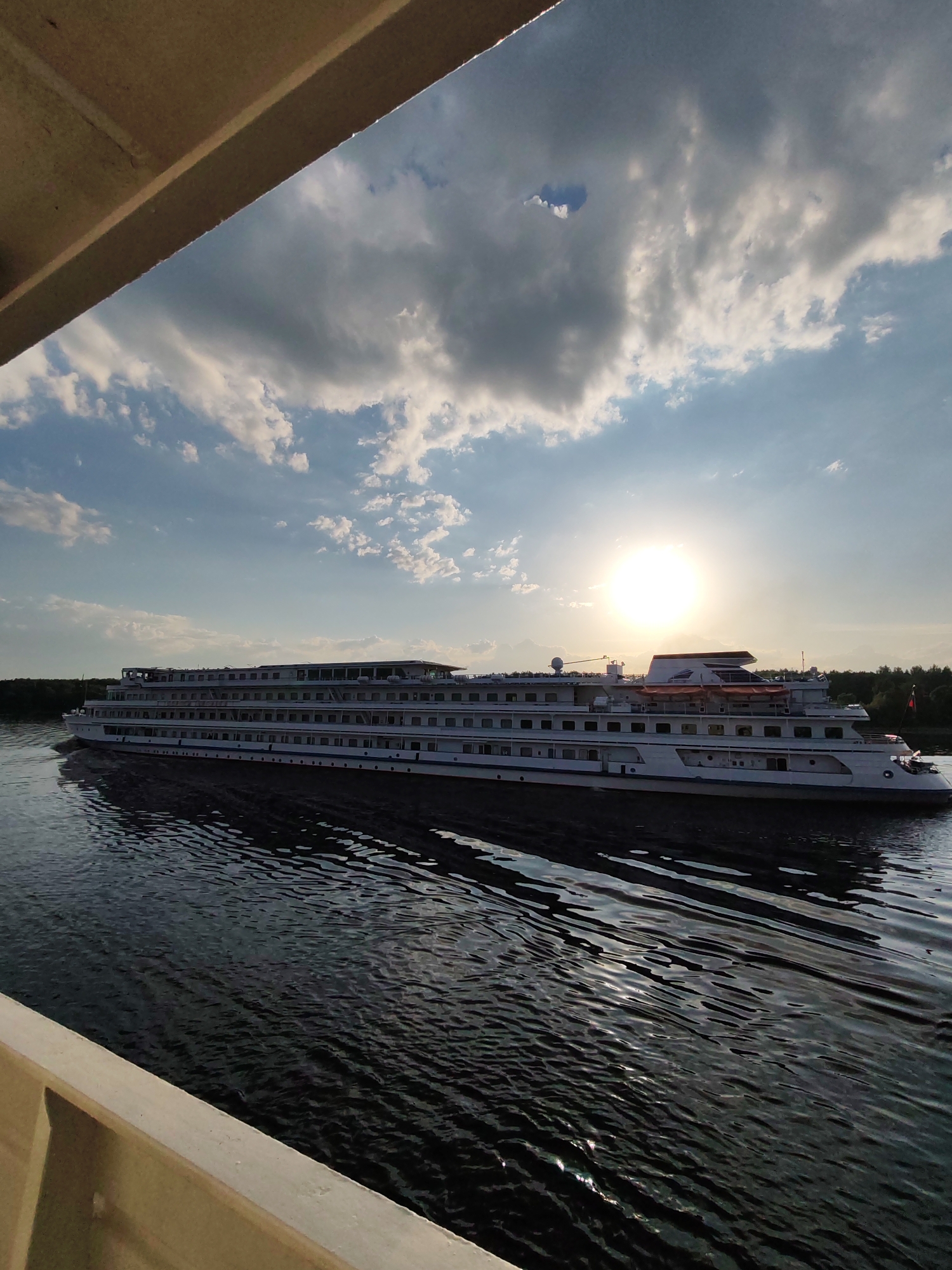 The image, taken in Novye Shatrishchi, Russia, depicts a multi-decked river cruise ship moving through the water. The ship is white with multiple rows of windows and appears to be a passenger vessel. The sun is visible through clouds, creating a bright spot in the sky. Dark, rippled water fills the foreground. Part of a structure, possibly the side or roof of another boat, is visible in the bottom left and top left corners of the image. Trees are visible along the shoreline in the background.