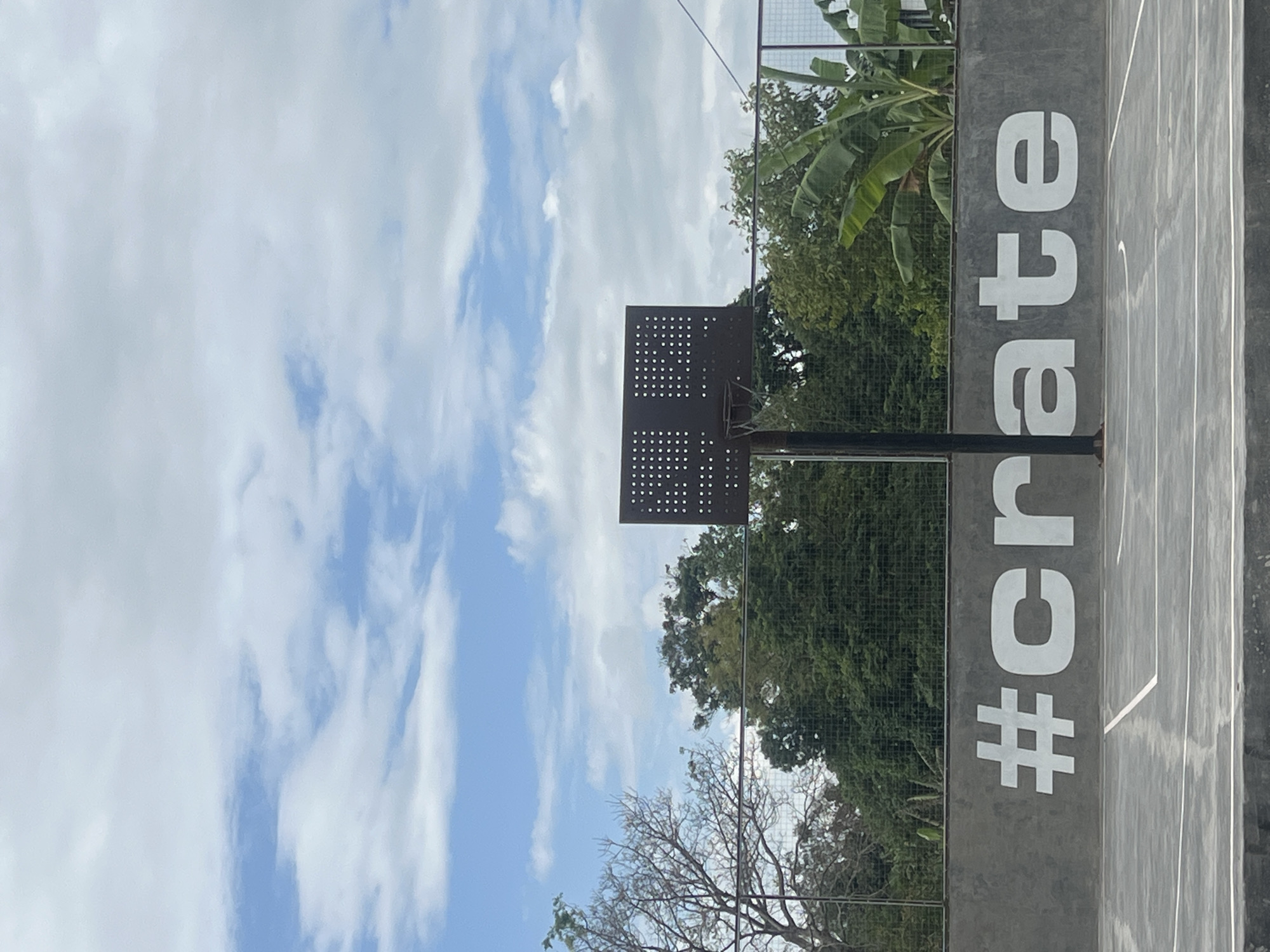 The image shows an outdoor basketball court in Mengwi, Indonesia. The court surface is grey with white lines. To the right, a concrete wall is visible with the word "#create" painted vertically in large white block letters. Behind the wall, trees and greenery can be seen, including what appear to be banana trees. A basketball hoop and backboard are positioned on the court. The sky is partly cloudy with blue sky peeking through white clouds, suggesting a daytime setting. The overall impression is of a well-maintained public space or recreational facility.