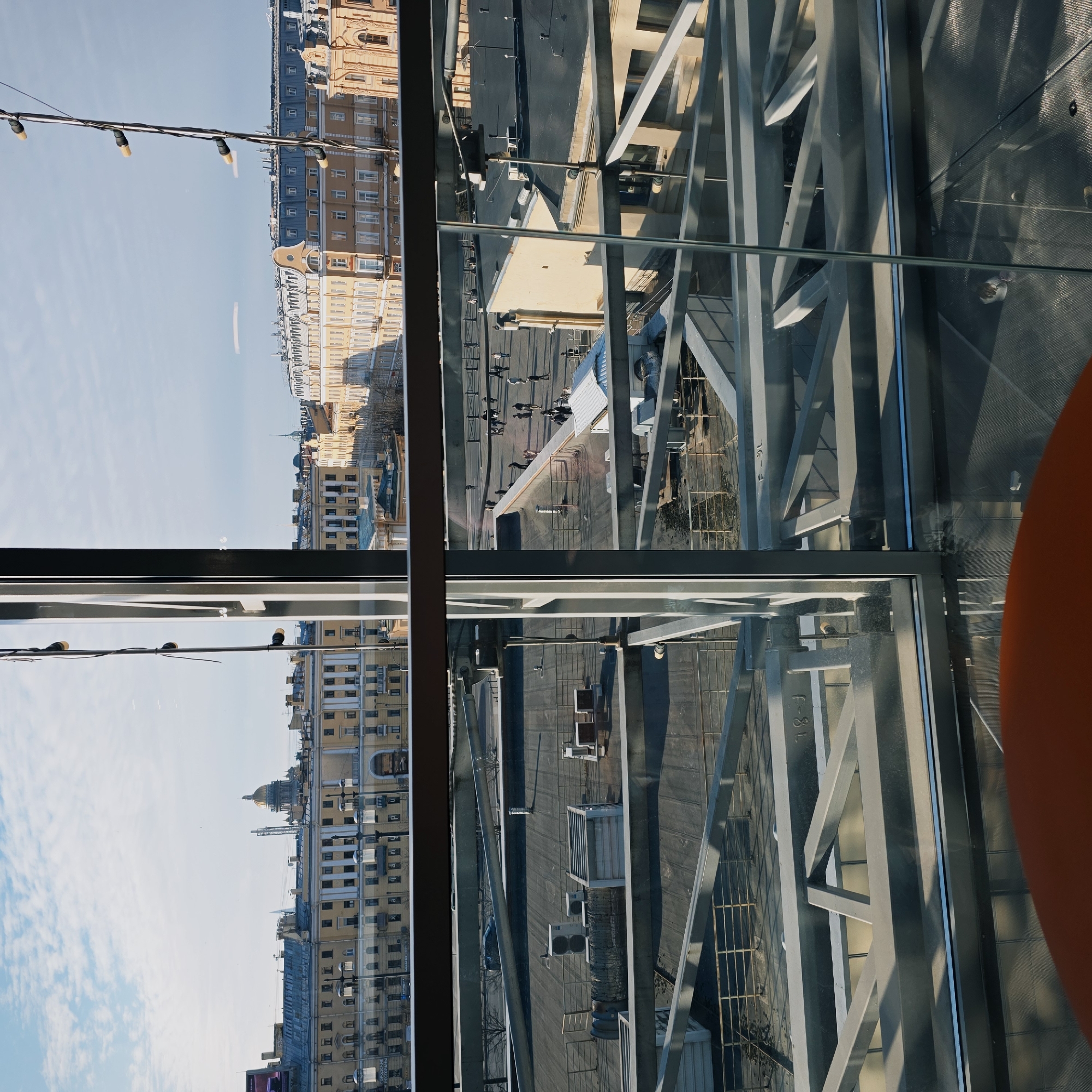 The image depicts a view through a window of a building in the city of Unknown, Unknown. In the distance, there are several buildings with a yellow-tan facade, some of which have architectural domes on top. The foreground showcases the architectural structure of the building itself, comprising metal beams and glass panes. Through these, there's a glimpse of a rooftop with ventilation systems and other mechanical fixtures. Below, people are seen walking on a pedestrian pathway. Additionally, some string lights are visible in front of the window.