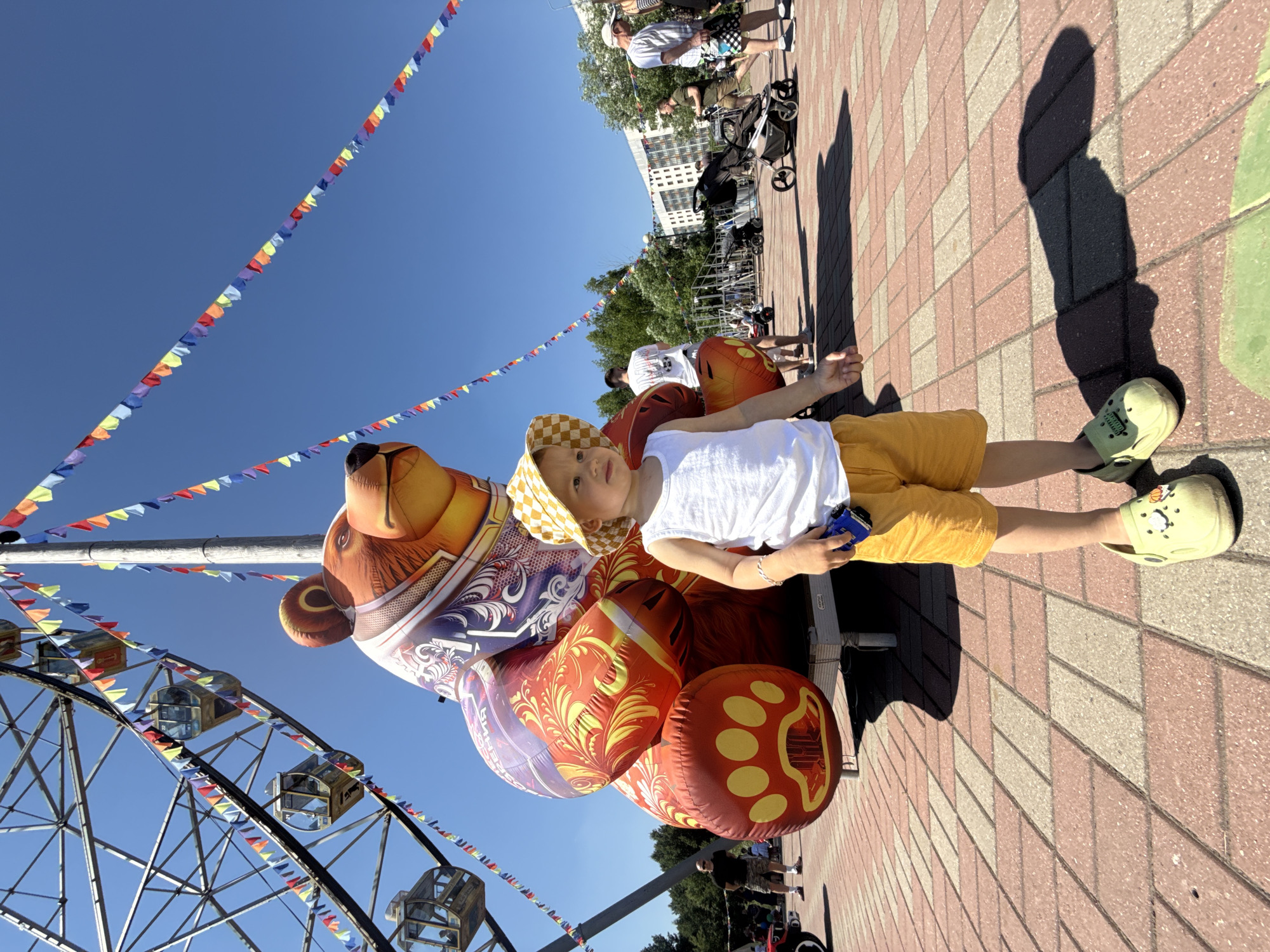 In the city of Zavolzhye, Russia, on a bright, sunny day, a young child stands in the foreground on a patterned brick walkway. The child is wearing a white tank top, yellow checkered bucket hat, and yellow shorts, holding a small blue toy car. To the left and slightly behind the child is a large, inflatable, cartoonish bear. The bear is orange and red with yellow accents and paw prints on its feet, and it wears a decorative purple and white patterned vest. Behind the bear, a Ferris wheel looms, adorned with colorful triangular flags strung on a pole. In the background, several adults and children are visible, along with strollers and buildings, suggesting a public gathering or park setting. The sky is a clear, vibrant blue.