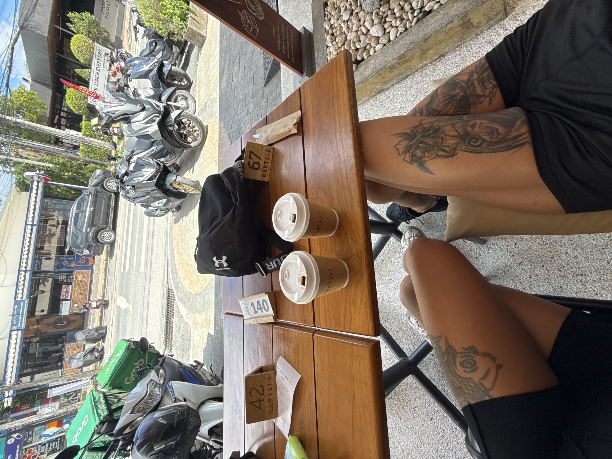 The image shows an outdoor seating area with two wooden tables. On the left table, there is a receipt and a green object, possibly a phone. A wooden table number sign displaying "42 BARTELS" is also present. On the right table, there are two paper cups with lids, a small paper packet, and a wooden table number sign displaying "67 BARTELS". A black backpack with the Under Armour logo is placed on the right table.
In the background, a street scene is visible with several motorcycles parked. Two delivery motorcycles with large green bags are prominent in the foreground. Further back, more scooters and cars are visible, along with buildings that appear to be shops or restaurants. The street has a pedestrian crossing. The ground in the foreground is paved with grey and patterned tiles, and a low wall with stones is visible on the right.
Two individuals are seated in the foreground, with their legs visible. The person on the left is wearing black shorts and white sneakers. Their left leg is visible with a tattoo of a stylized face on the thigh. The person on the right is wearing black shorts and black sneakers. Their legs are visible, with tattoos of a rose and a face on the right thigh and a design on the left thigh. The overall scene suggests a casual outdoor cafe or dining setting in Choeng Thale, Thailand, likely a popular area for both locals and tourists, given the presence of delivery services and various shops.