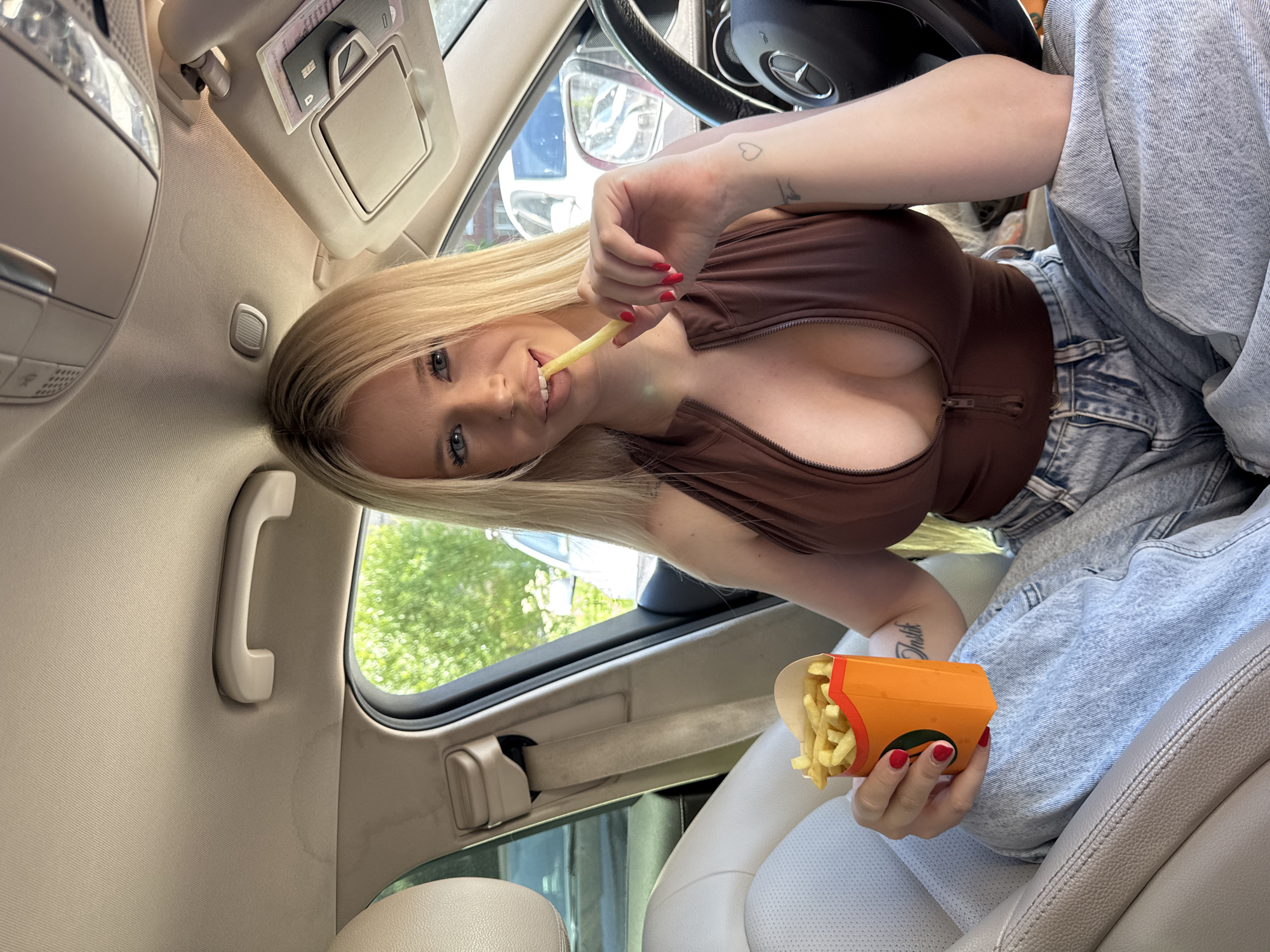 A blonde woman with blue eyes is sitting in the driver's seat of a car and eating french fries. She is wearing a brown zipped top and blue jeans. The car is parked on a street in Saint Petersburg, Russia, with trees and buildings visible outside the windows. The woman is holding a box of french fries in her left hand and is eating a french fry with her right hand. She is looking at the camera and smiling.