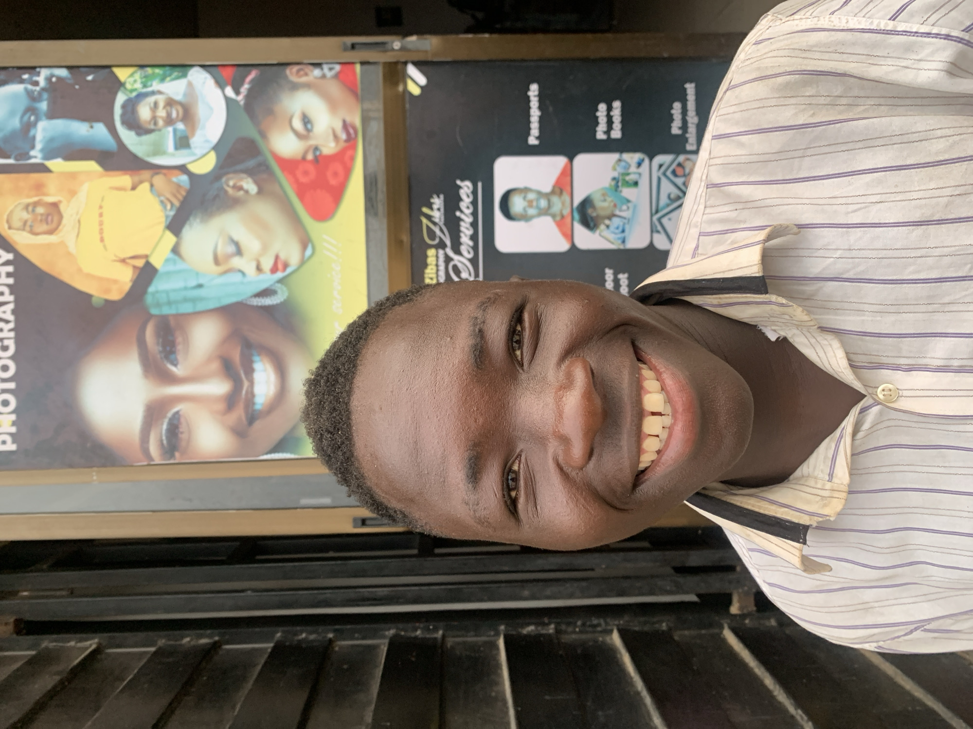 A close-up, slightly angled shot depicts a smiling dark-skinned man wearing a striped shirt. The photo is taken in front of a photography services storefront, “Zibas Zlat Services” with signage advertising “Photography”, “Passports”, “Photo Books”, and “Photo Enlargement”. The background features photographic portraits on display. The location is potentially in Jos, Nigeria, based on contextual information.