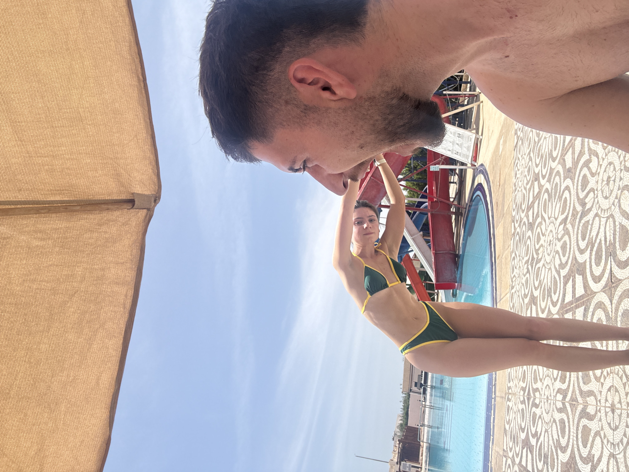 The image depicts a man and a woman in an outdoor setting near a swimming pool. The man, who appears to be in the foreground, is partially visible with his head and upper body filling the top of the frame. He has short dark hair and a beard. The woman, who is further away, is wearing a green and yellow bikini and has her arms raised. She is standing on a patterned tiled surface next to a pool of water. In the background, there is a water slide and buildings, suggesting a resort or water park environment. The sky is blue with some clouds visible. An umbrella is seen to the left of the picture. The city is Unknown, Unknown.