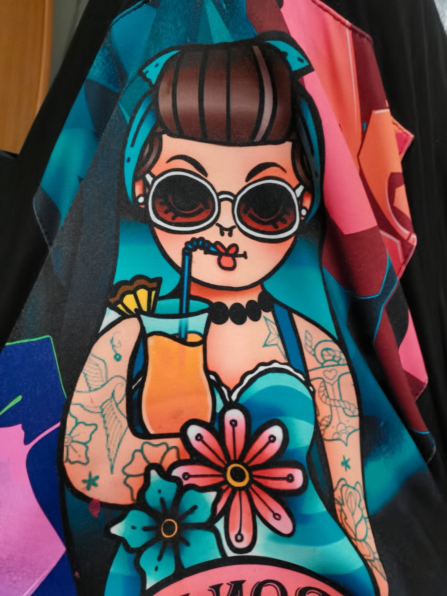The image displays a vibrant, close-up photograph of an illustration printed on a piece of clothing, likely a T-shirt or similar garment, with visible fabric folds and texture. The central subject is a stylized cartoon woman with a distinct retro or pin-up aesthetic. She has brown hair with prominent blue streaks, pulled back with a matching blue headband. Large, round, white-framed sunglasses obscure her eyes, and her red lips are slightly pursed around a blue straw. A black beaded necklace adorns her neck.
She is dressed in a blue top or dress with a sweetheart neckline, from which colorful flowers, including a large pink one with a yellow center and a smaller blue one, appear to emerge. Her arms are heavily adorned with outlines of various tattoos, such as a bird, flowers, an anchor, a star, and a heart with a rope motif. She holds a tall cocktail glass filled with an orange beverage, garnished with a yellow fruit wedge.
The background of the illustration features abstract geometric patterns in shades of blue, pink, orange, and burgundy, consistent with the overall design. Partial text is visible at the bottom of the image, appearing to spell out "GOOD VI". The woman's confident posture and the cocktail suggest a relaxed or celebratory mood. This media file was taken in Chaikovsky, Russia.