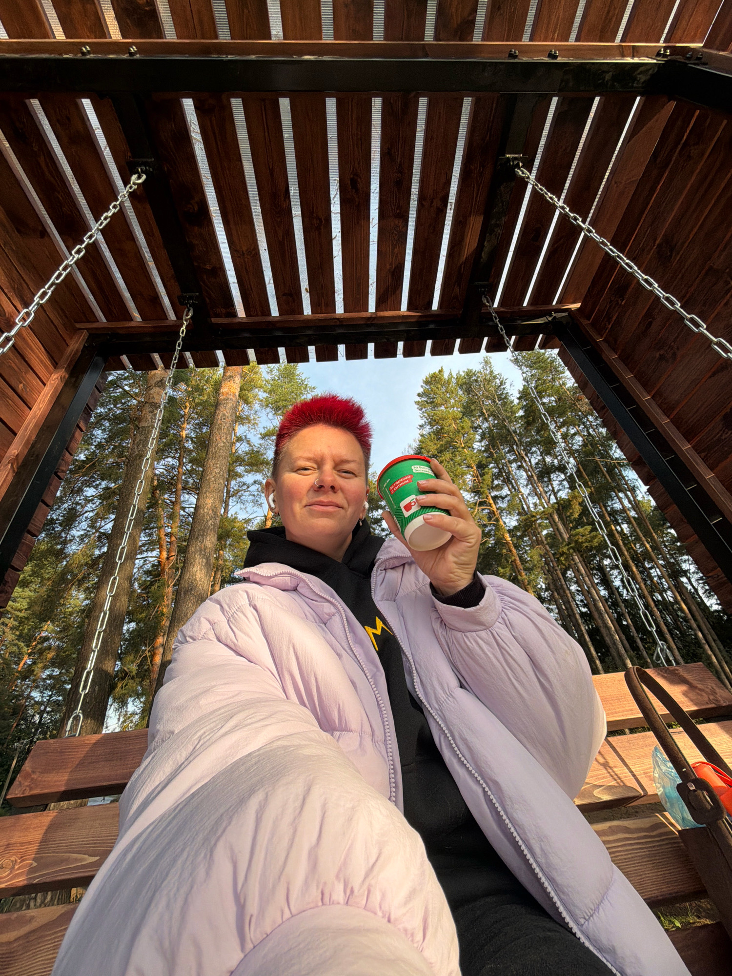 A low-angle, wide shot captures a person with bright red hair sitting on a wooden swing under a slatted wooden roof. The person is holding a green and white paper cup in their right hand, which is raised towards the camera. They are wearing a light purple puffer jacket over a black hoodie, and white earbuds are visible in their ears. The swing is suspended by metal chains from the roof structure. Behind the person, a dense forest of tall pine trees is visible, suggesting an outdoor, natural setting. The sun appears to be shining, casting light through the trees and onto the swing. A brown tote bag with some items peeking out is visible to the right of the swing. The scene is consistent with a park or recreational area in Chaikovsky, Russia, given the prevalence of pine forests.