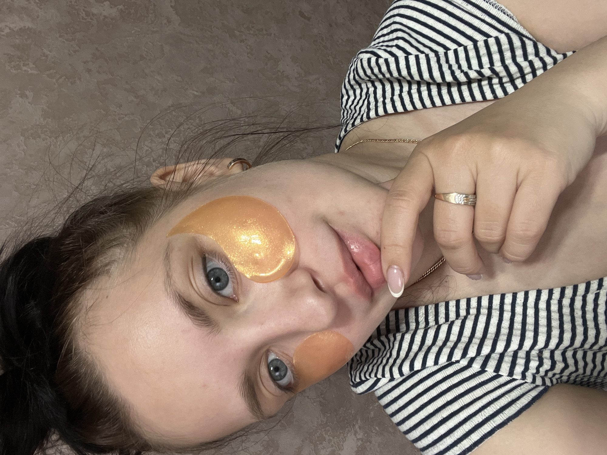 A woman lies on a gray-brown surface, likely a bed or floor, wearing a black and white striped shirt. She has light skin, dark hair, and blue eyes. On her face, she wears two golden eye patches under her eyes. She has a ring on her finger, and her hand is near her mouth. The location is Марковский, Russia.