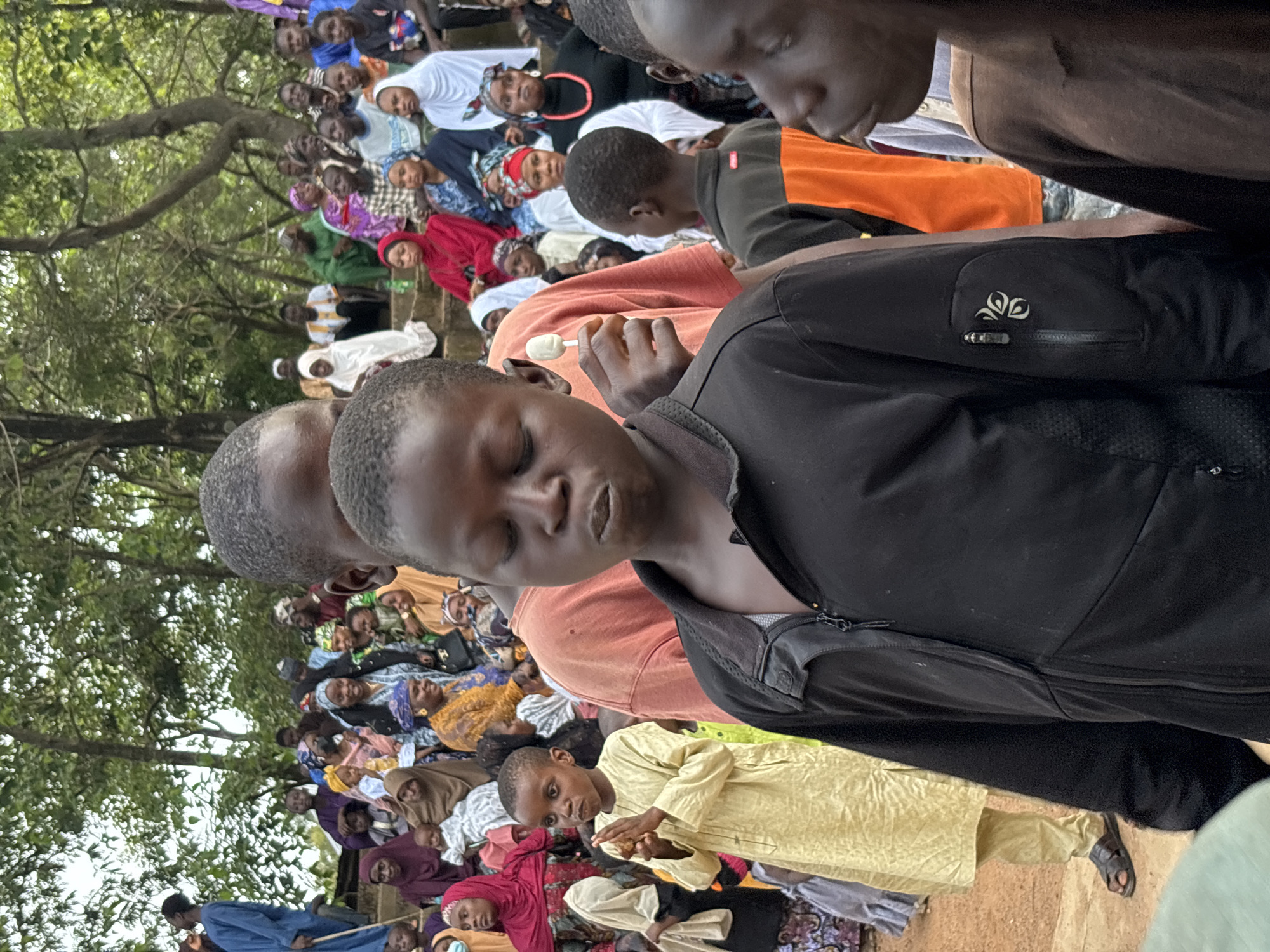This is a medium shot photograph taken outdoors, likely in an urban environment in Jos, Nigeria, during the daytime. The scene is populated with a crowd of people, primarily children and adults, dressed in a variety of traditional and modern clothing, many wearing head coverings. The background features trees and steps made of stone or concrete, suggesting an outdoor gathering space.
In the foreground, a young boy in a black zip-up jacket with a distinct logo on the pocket is the closest subject. His eyes are closed, and his mouth is slightly open. To his left, another young boy wearing a light yellow traditional garment is holding his hands together and looking forward. His face is clearly visible. Behind these two boys, an adult wearing an orange t-shirt is visible from the neck down. Further back, other individuals are observed in the crowd, some are holding children. The overall atmosphere appears to be a community gathering, possibly an event or a religious ceremony, given the attire of many of the individuals. The ground appears to be a reddish-brown soil.