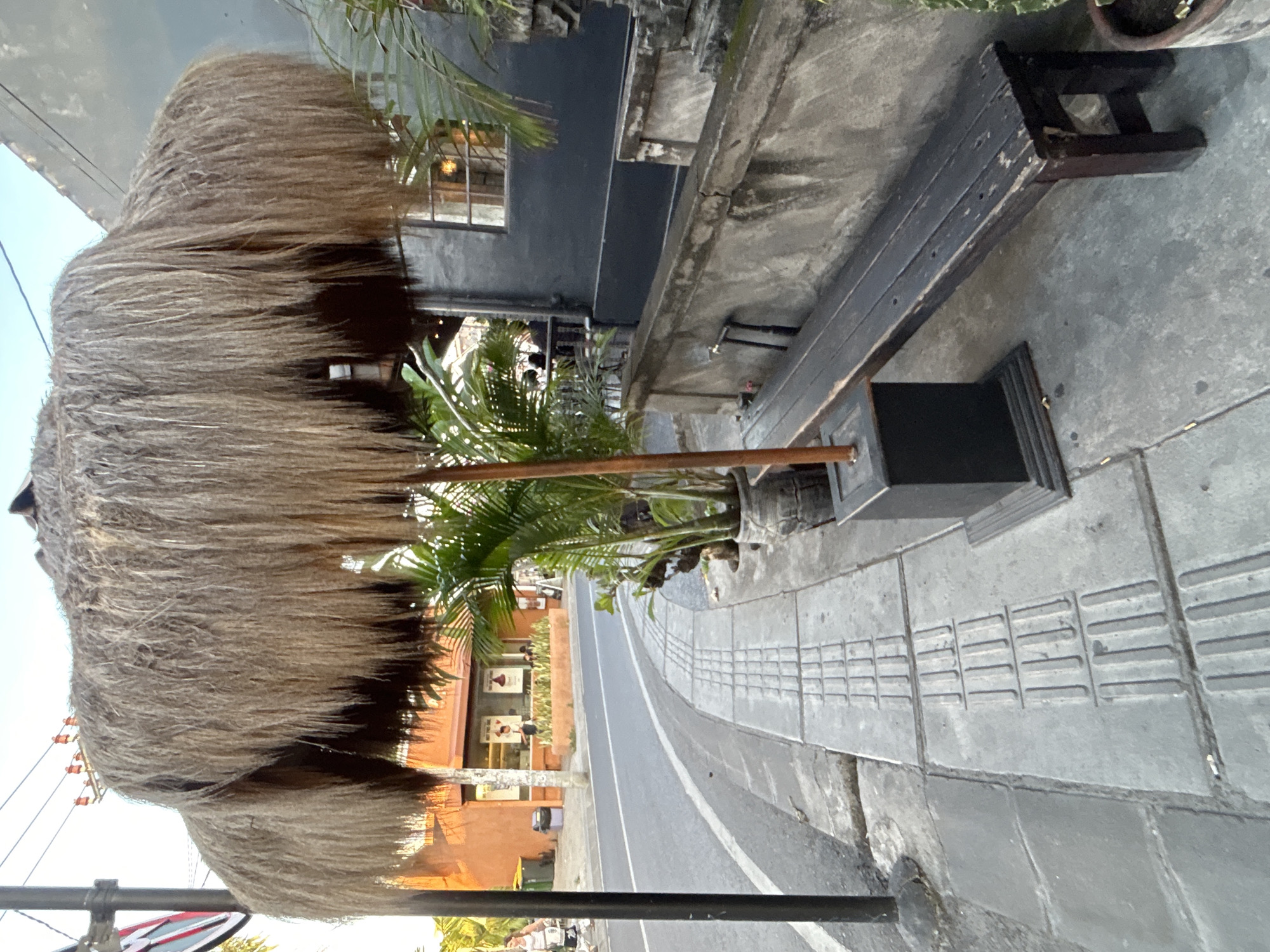 The image captures a street scene in North Kuta, Indonesia, focusing on an outdoor seating area next to a road. A large, thatched umbrella provides shade over a dark wooden bench positioned along a concrete sidewalk. The sidewalk features tactile paving, indicating accessibility features. In the background, an orange building with shop windows and an awning is visible, and a few palm plants are placed in pots near the bench. The road curves away to the left, with a white line marking the edge. Power lines are visible against the sky. A dark lamppost stands on the left side of the frame. The overall impression is that of a relaxed, possibly commercial, outdoor space in a tropical urban setting.