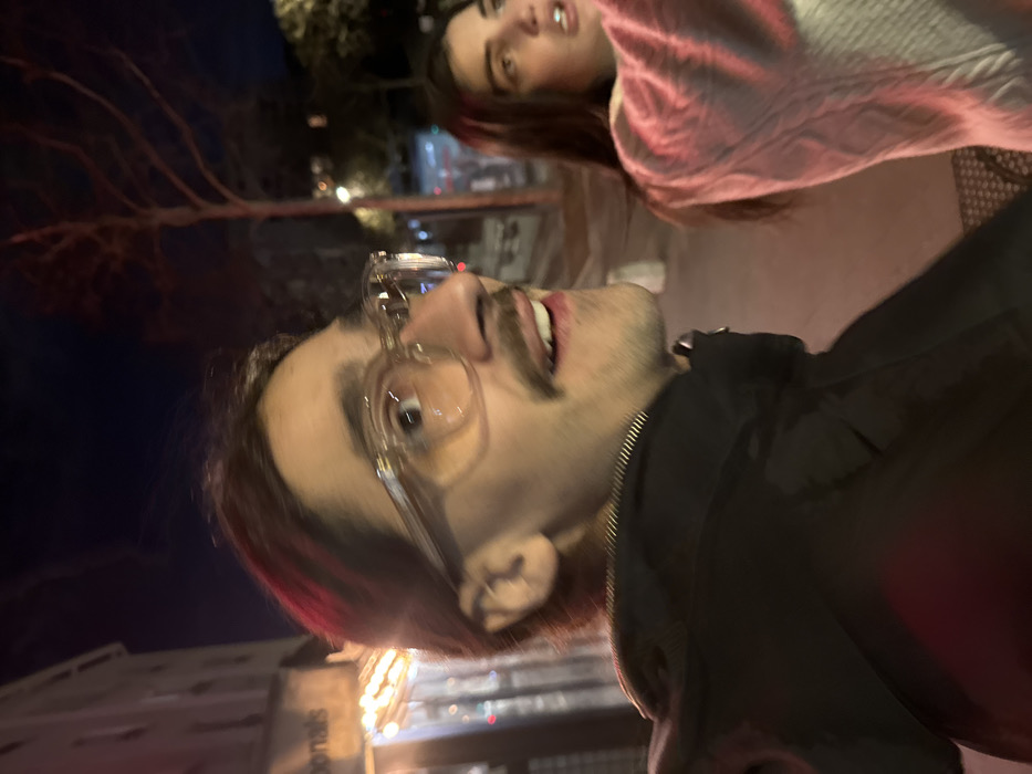 The image depicts a nighttime urban scene in Vincennes, France, featuring two individuals outdoors. In the foreground, a male with dark, pulled-back hair, a mustache, and a goatee wears clear-framed glasses and a black zippered jacket. He is looking sharply to his right and upward, with his mouth slightly open. Behind and to his right, a female with long dark hair is partially visible, wearing a light-colored cable-knit sweater with red illumination on her left arm. She is also looking upward and to her right, with her mouth slightly open.
In the background on the left, a light-colored building with multiple stories is visible. An awning or sign structure displays partially visible letters "onalds," suggesting a McDonald's establishment. Below this, warm-toned string lights are strung horizontally across the building's exterior. Further back and to the right, a bare-branched tree stands against the dark night sky, with additional blurred lights and distant foliage visible. The ground appears to be a paved surface or sidewalk, with a textured element resembling outdoor seating or a grate visible in the lower right.