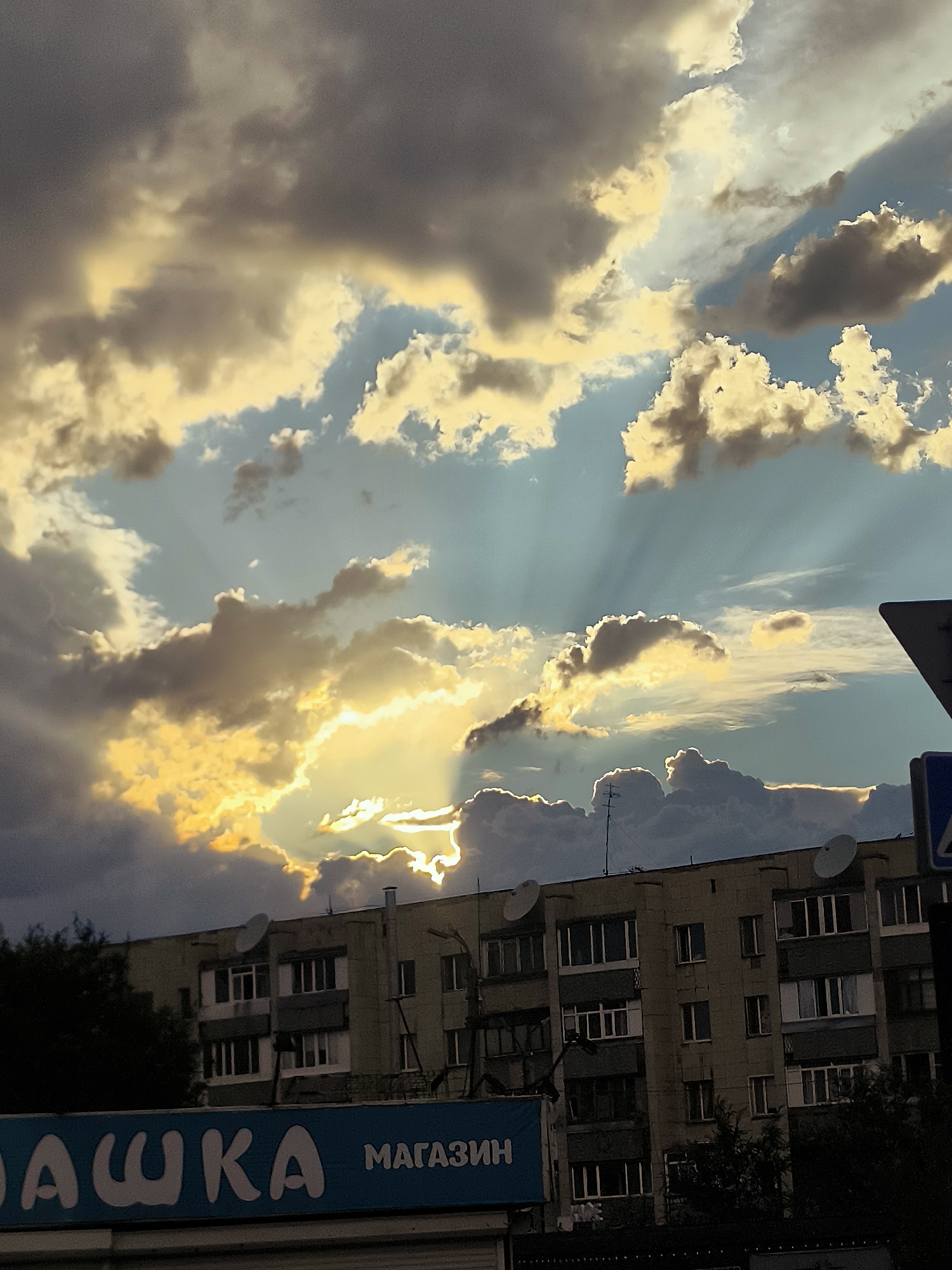 The image captures a dramatic sky over a cityscape. The sky is filled with a mix of dark, brooding clouds and brighter, sunlit clouds, with rays of sunlight piercing through. Below the sky, a multi-story apartment building is visible, its facade showing a grid of windows. To the left and bottom of the image, a blue sign with white lettering is partially visible. Trees are present on the left side of the building and also in the foreground. A street sign is visible on the right side of the image. The overall impression is of a dynamic and somewhat moody atmosphere, possibly at sunset or sunrise, in an urban environment.