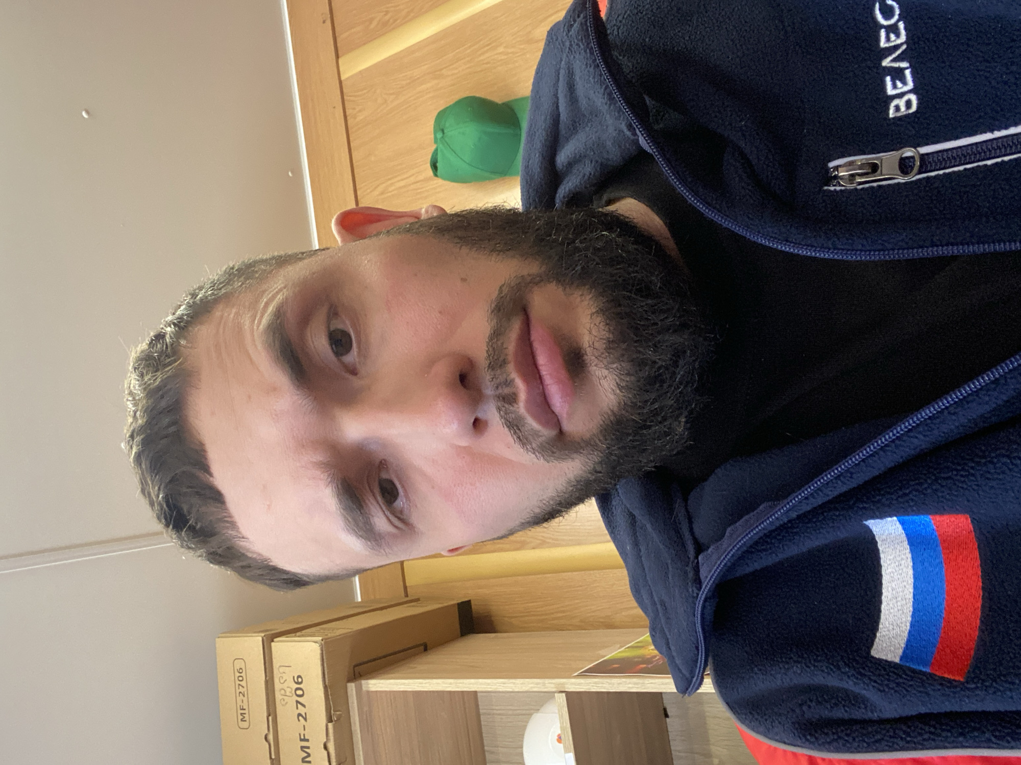 The image displays a man taking a selfie or being photographed indoors. He is positioned in the foreground, looking directly at the camera with a neutral expression. The man has dark hair, styled short on the sides, and a full dark beard.
He is wearing a dark blue fleece jacket with a zipper running down the front. A dark undershirt or turtleneck is visible beneath the jacket. On the right side of his chest, near the zipper, white embroidered Cyrillic text spells "ВЕЛЕС" (VELES). On his left sleeve, there is an embroidered rectangular patch featuring the Russian flag, characterized by horizontal stripes of white, blue, and red.
The background consists of light-colored wooden walls and shelving units, indicating an indoor setting. On a shelf to the man's lower left, two cardboard boxes are visible, one prominently displaying the text "MF-2706" and what appears to be "ЭВЭН" (EVEN) in Cyrillic. A green baseball cap rests on a shelf behind him. Another white, possibly ceramic, object is situated on a lower shelf. The lighting is consistent with an indoor environment. The image provides no specific cues for the time of day or weather. This scene is contextualized as taking place in Luzhitsy, Russia.