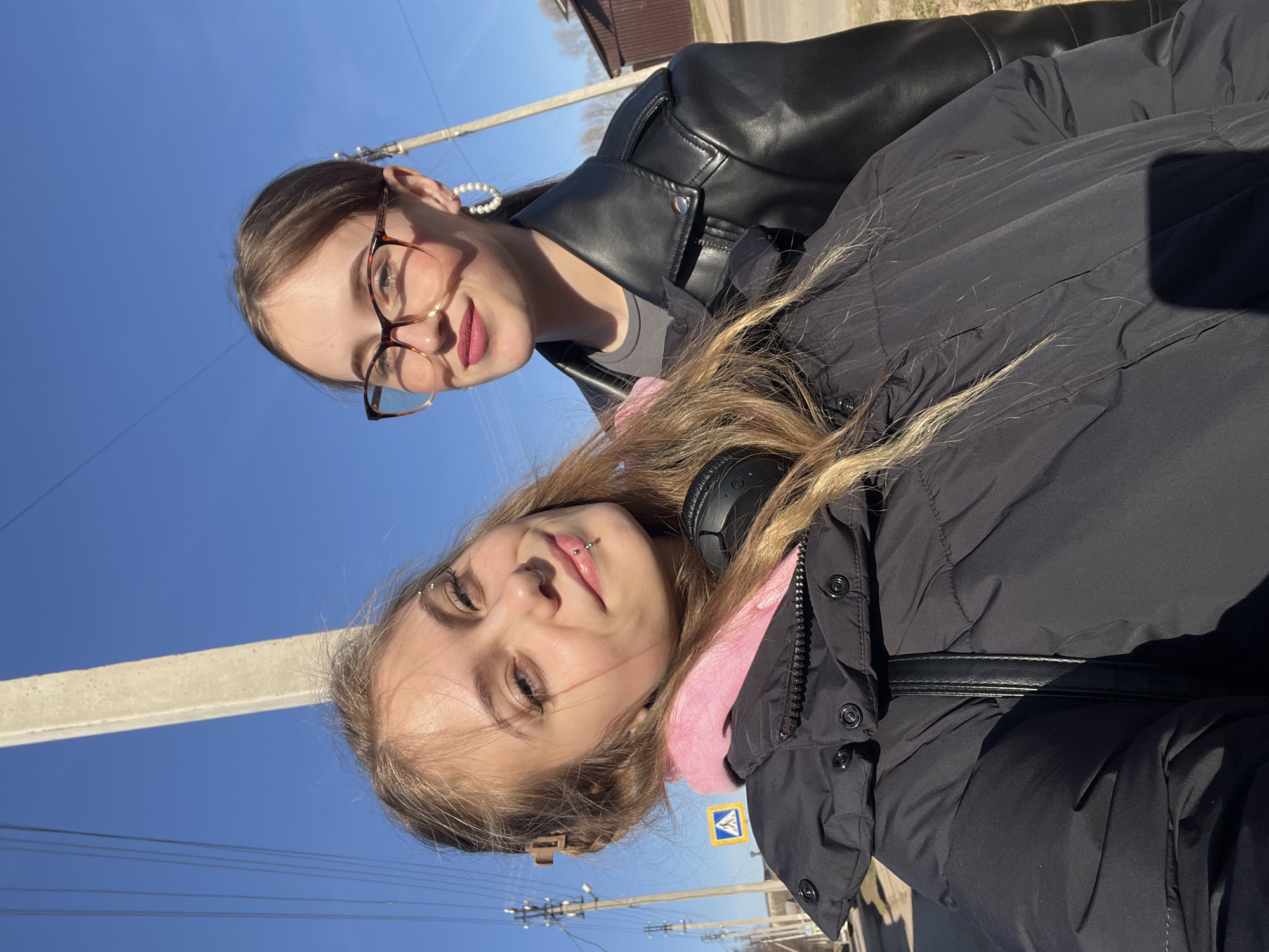The image features two young women posing for a selfie outdoors in Фоки, Russia, on a clear, sunny day, likely midday or early afternoon given the bright lighting.
**Subjects:**
The woman in the foreground has blonde hair, is wearing a black puffer jacket over a pink top, and black over-ear headphones around her neck. She has several facial piercings including a labret, an eyebrow piercing, and a nostril piercing. The second woman, positioned behind her, has brown hair, wears brown-framed glasses and large pearl hoop earrings, and is dressed in a black leather jacket over a grey top.
**Activity or event taking place:**
The two women appear to be posing for a casual selfie, both looking towards the camera with gentle, relaxed smiles.
**Notable details:**
The sky is a bright, clear blue, indicating excellent weather. Power lines and a concrete pole are visible in the background against the sky. A yellow and blue road sign, clearly displaying a pedestrian crossing symbol, is visible near the bottom right. Distant buildings suggest an urban or semi-urban environment.
**Visible text:**
The most prominent visible text is the symbol for a pedestrian crossing on the road sign. No discernible alphabetic text is clearly visible.