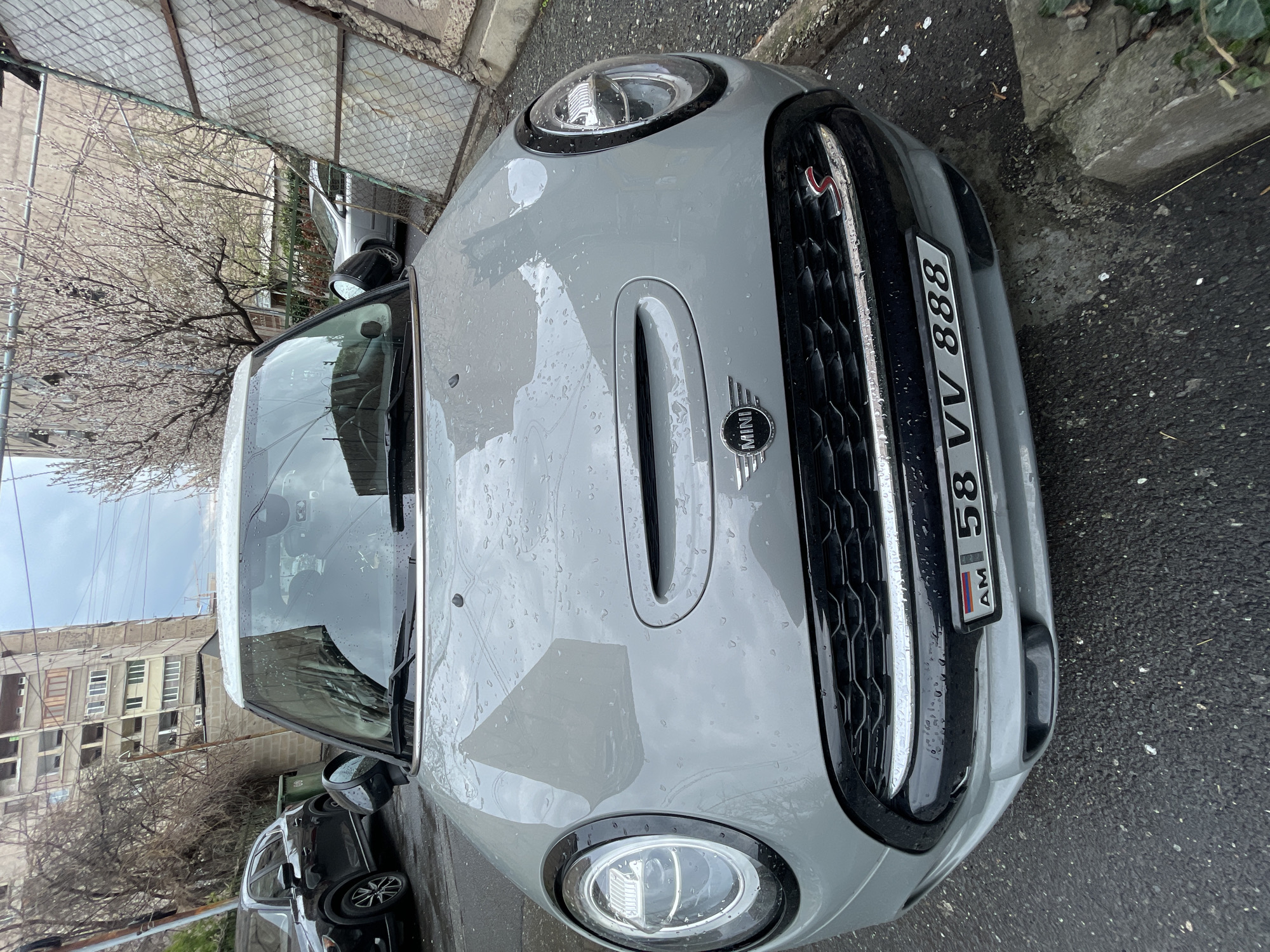 The image shows the front of a gray Mini Cooper parked on a wet street. The license plate reads "AM 58 VV 888." The car is parked on what appears to be a city street in Yerevan, Armenia, as indicated by the "AM" license plate. A building is visible in the background, along with a bare tree and a fence. Another car is partially visible next to the Mini Cooper. It's a cloudy day, and the car's hood and windshield are covered in raindrops.