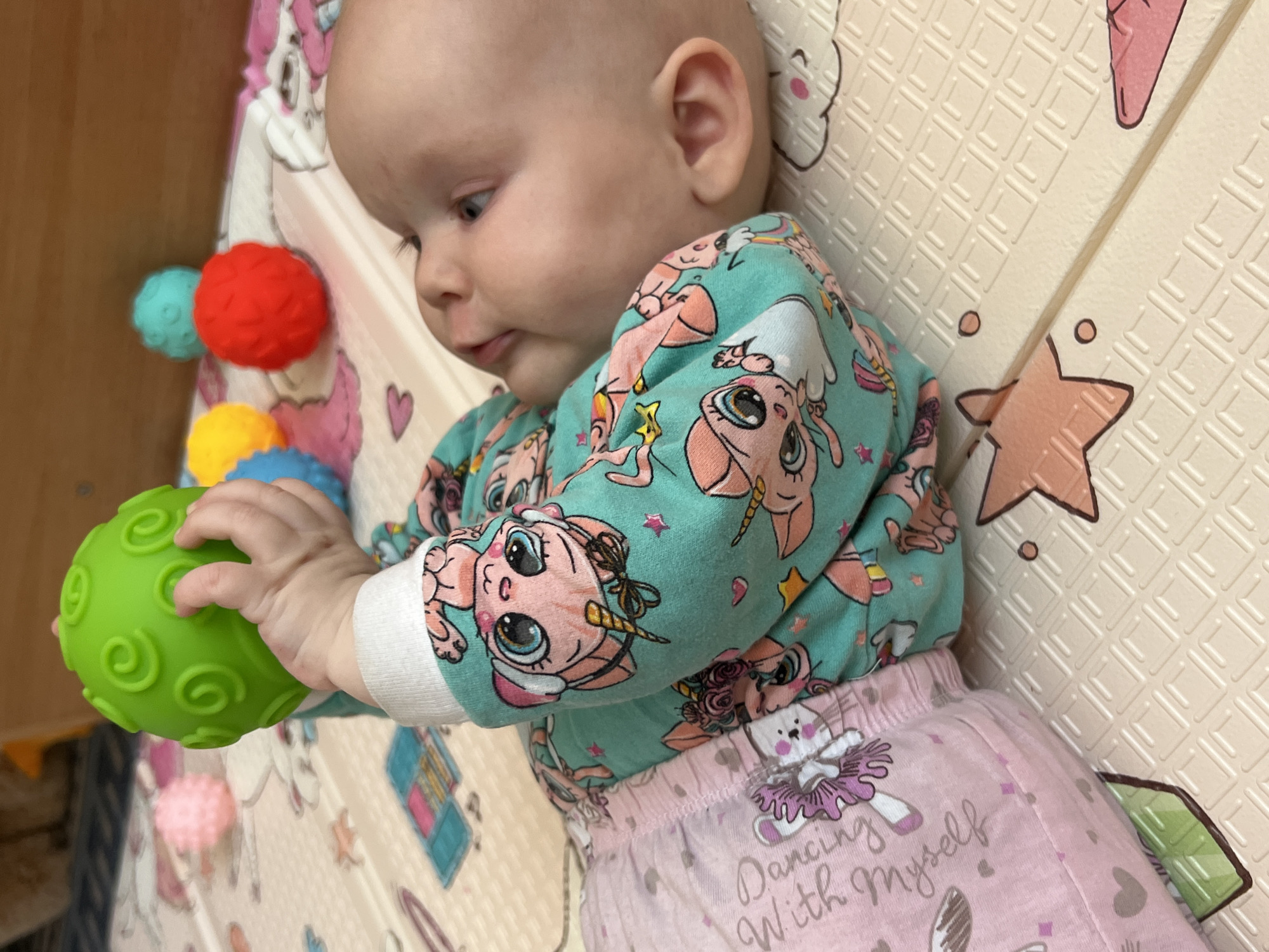 A baby lies on a beige padded play mat with pink star and cloud designs. The baby is wearing a turquoise long-sleeved shirt with pink cartoon unicorn designs and pink pants that say "Dancing with myself" next to a ballerina bunny design. The baby is holding a green textured ball with both hands. Three colorful spiky balls, red, turquoise, and yellow, are near the baby. The setting appears to be indoors. The location of Votkinsk, Russia is not discernible from the image.