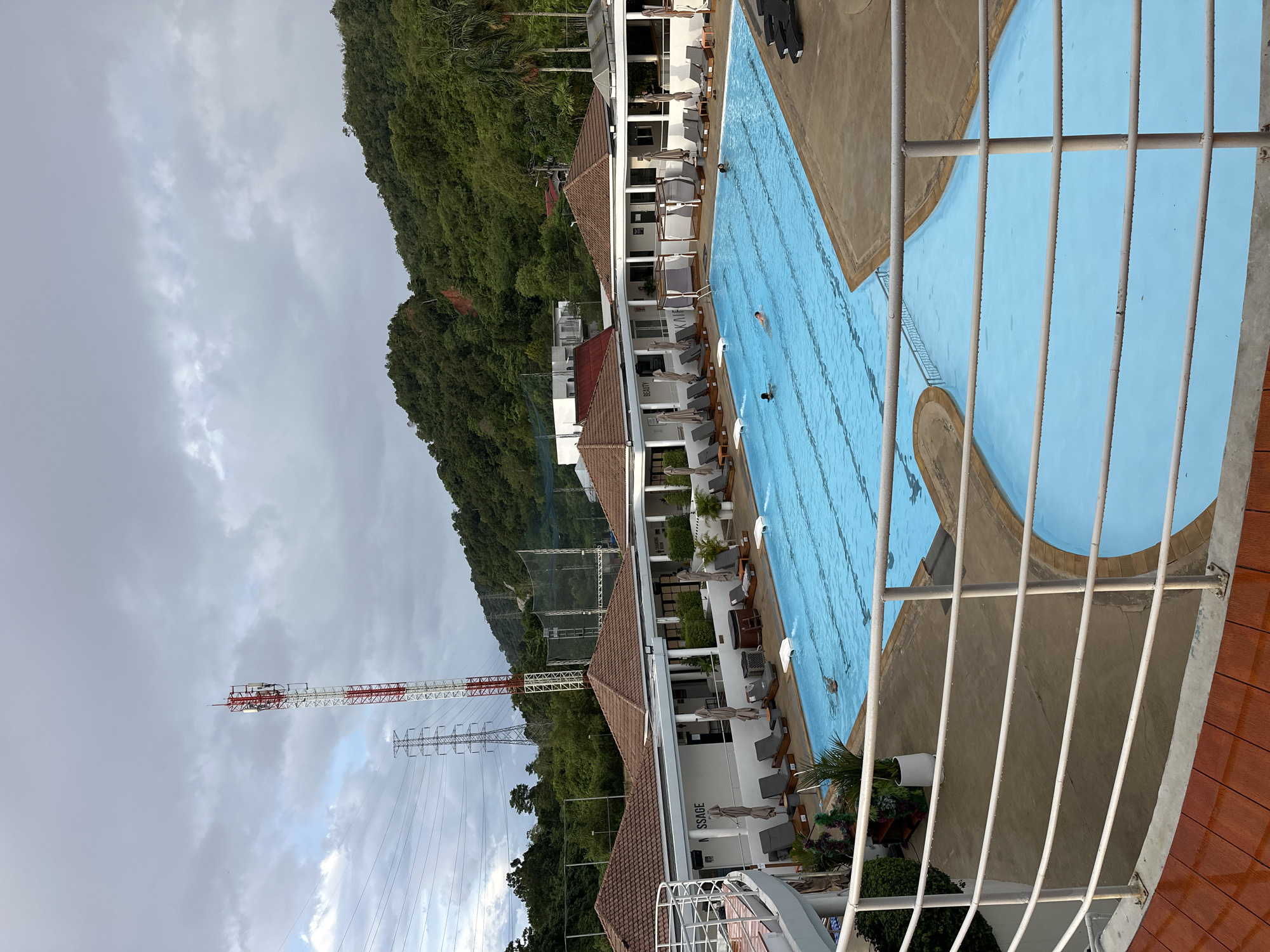 This image captures an outdoor swimming pool area at a resort or club in Kathu, Thailand. The pool is a bright turquoise blue, with lane markers visible in the larger section. Several lounge chairs with umbrellas are set up around the pool, and some are occupied by people relaxing. A few individuals are swimming in the pool.
In the background, there are several white buildings with brown tiled roofs, suggesting cabanas or changing rooms. Some of these buildings have signage visible, including "MASSAGE" and "BEAUTY". A small hot tub is located next to the main pool.
The environment is lush and green, with dense tropical vegetation covering the hills in the background. A tall, red and white telecommunications tower is a prominent feature on one of the hills, along with high-voltage power lines and pylons. The sky is overcast with dramatic clouds, indicating potentially stormy weather. The foreground shows a white metal railing and a wooden deck area.