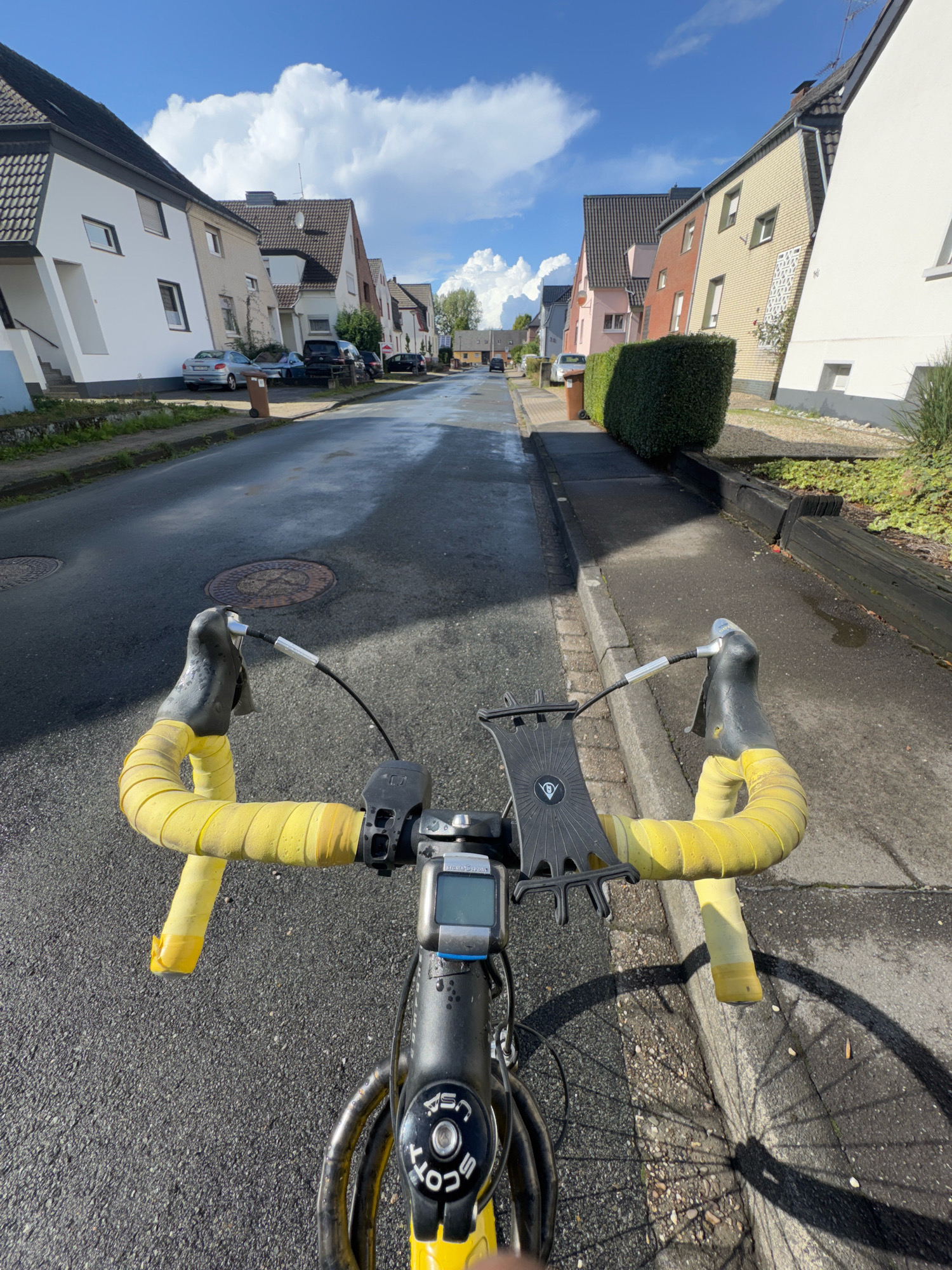 The image is a first-person perspective shot from a bicycle's handlebars, looking down a wet asphalt road lined with two-story houses. The sky is blue with scattered white clouds. The houses appear to be suburban residences, with light-colored exteriors and dark tiled roofs. There are parked cars on the side of the road. The bicycle handlebars are wrapped in bright yellow tape, and a black phone mount is attached. A small digital display is visible on the front of the bicycle stem. The road surface shows reflections from the sky and surrounding environment, indicating it is wet. The environment is a residential street, likely in Cleves, Germany, given the architectural style of the houses.