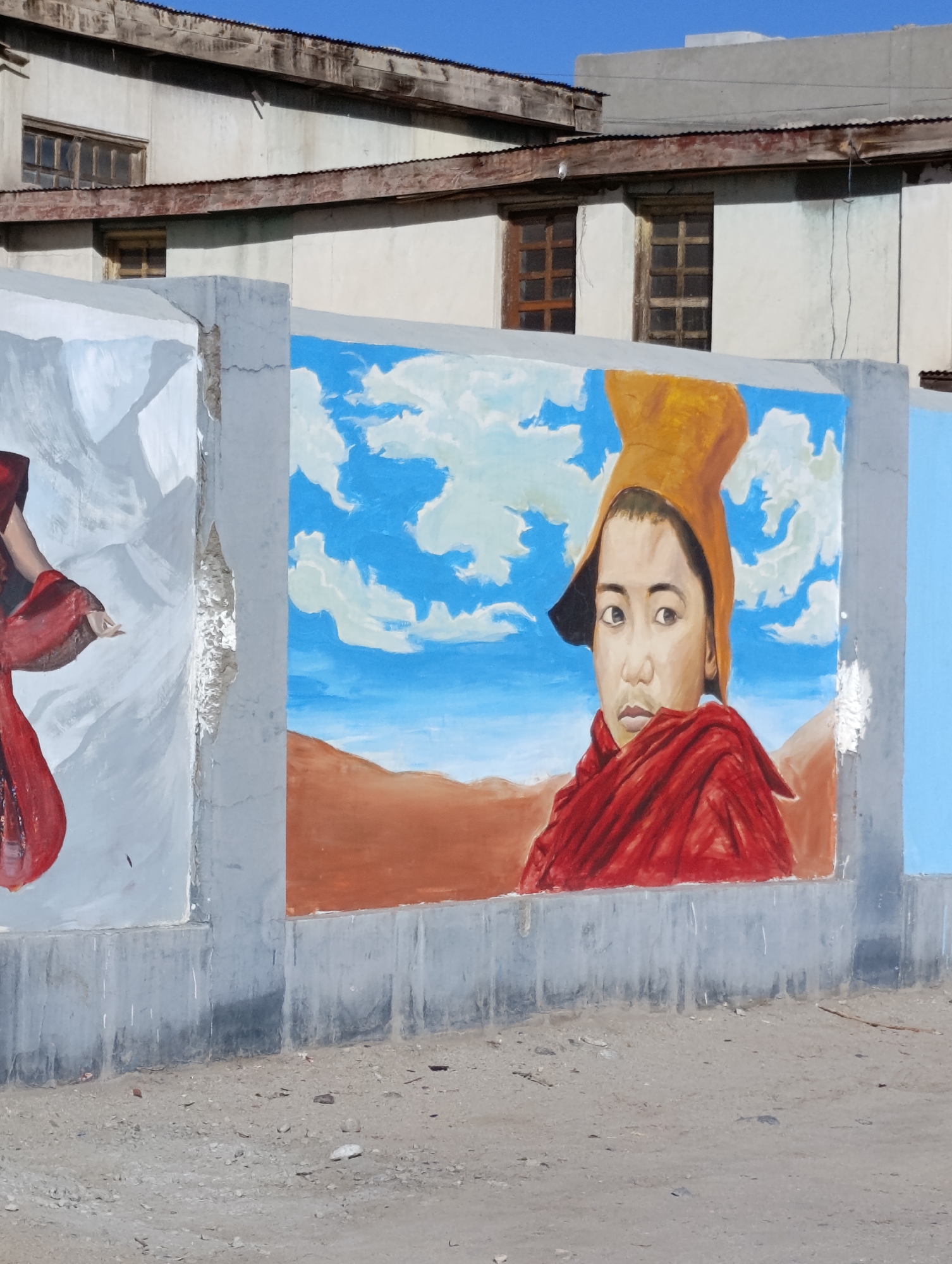 The image shows a mural painted on a concrete wall in the city of Leh, India. The mural depicts a young person wearing a yellow hat and a red robe, with a landscape of blue sky with white clouds and brown mountains in the background. To the left of this central figure, there is another partially visible figure painted in shades of white and red. The wall itself is gray and shows signs of wear and tear, with some cracks and peeling paint. Behind the wall, a building with a corrugated metal roof and windows can be seen. The ground in front of the wall is covered with gravel and dirt.