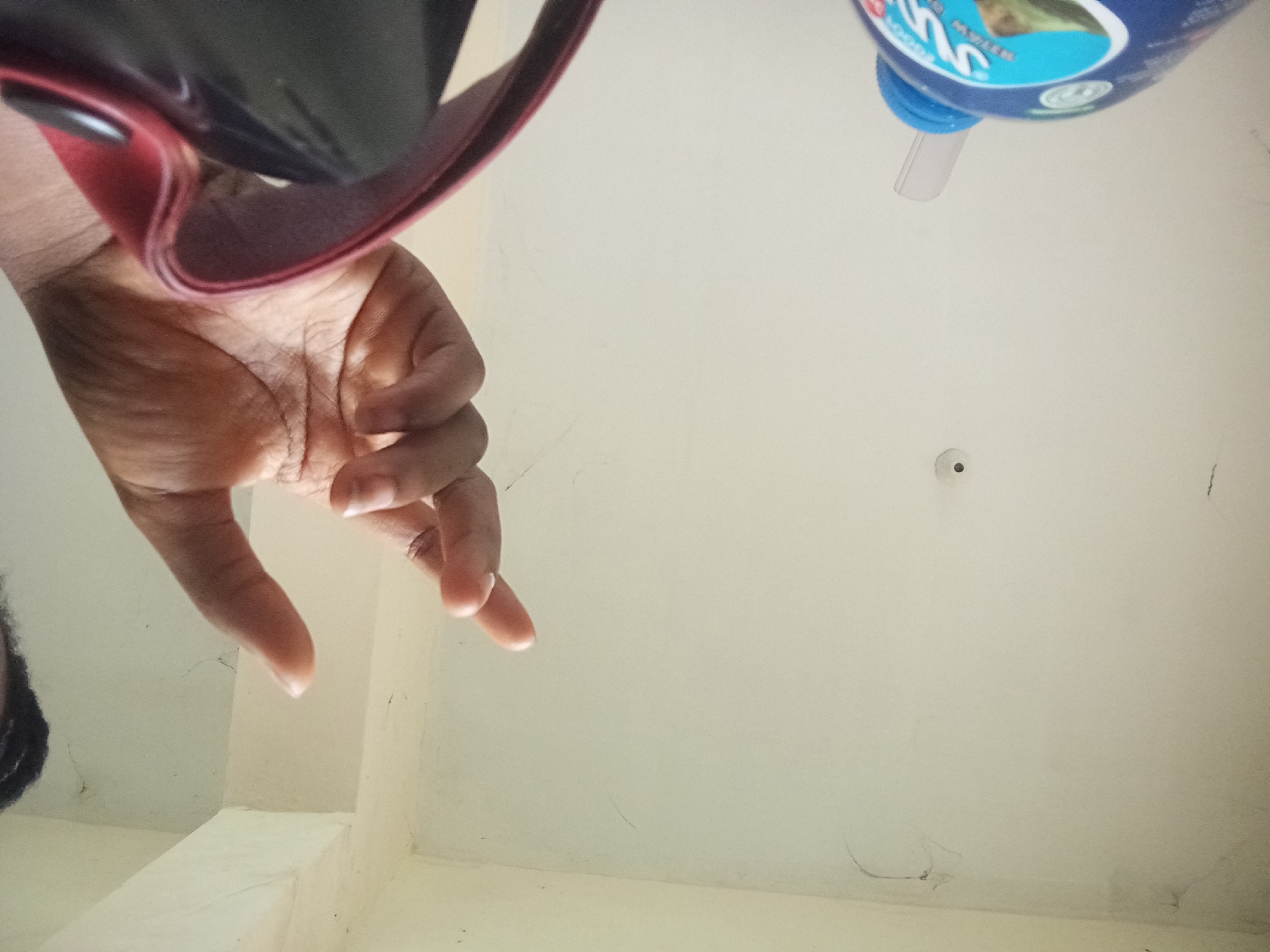 A low-angle, slightly blurry, indoor shot shows a person's hand reaching up towards a white ceiling. A portion of a blue plastic bottle with a label and a white cap is visible in the upper left corner. The hand is positioned in the center of the frame, with fingers slightly curled as if reaching for something. A red-lined bag or strap is partially visible in the lower left of the image. The ceiling appears to have a small circular object mounted on it, possibly a smoke detector or light fixture. There are some faint dark marks on the ceiling, suggesting dust or discoloration. The lighting is natural and diffuse, indicating an overcast day or indoor lighting.