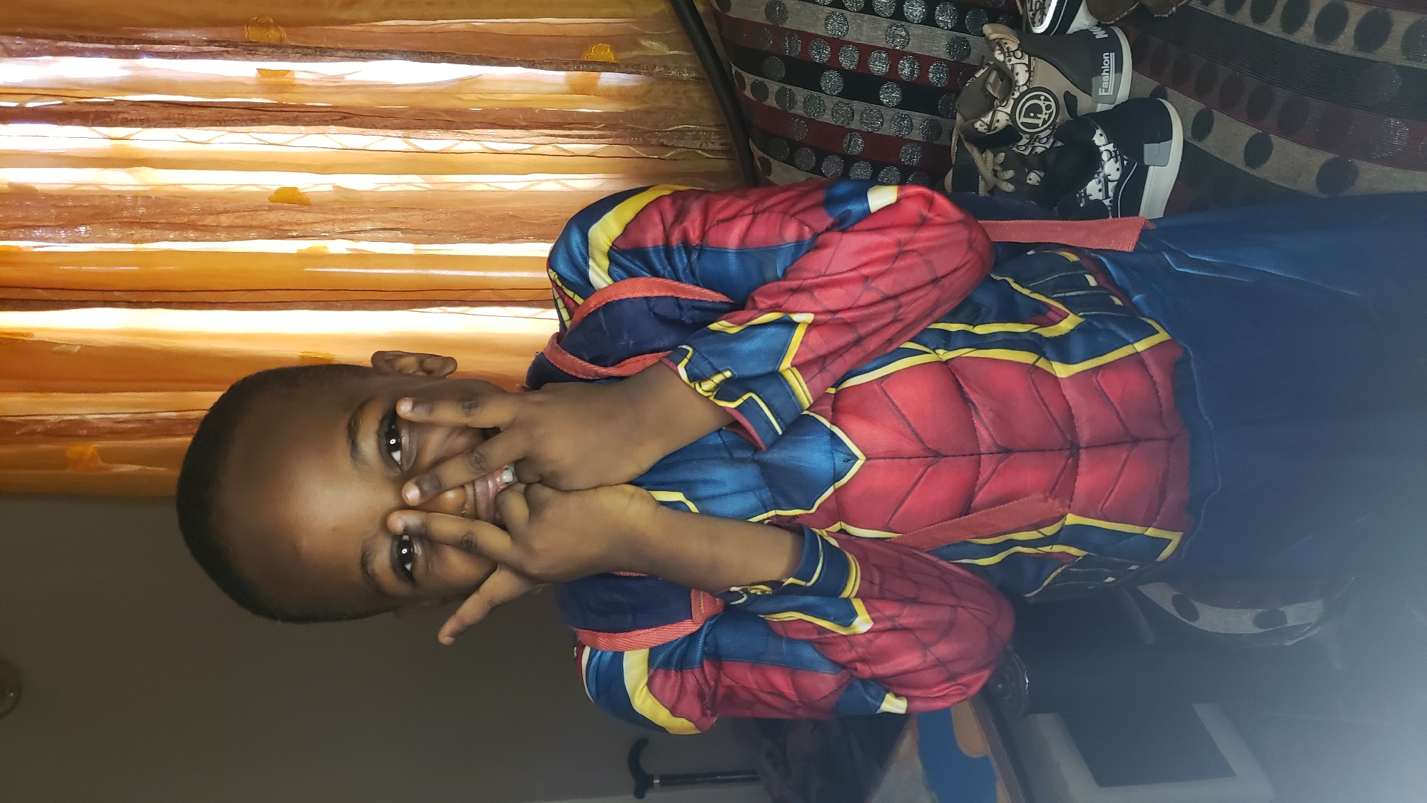 A young boy is posing in a Spider-Man costume. He is holding his hands up to his face and smiling. The background includes a window with sheer orange curtains, a patterned sofa, and a walking cane. The location is Narayi, Nigeria.