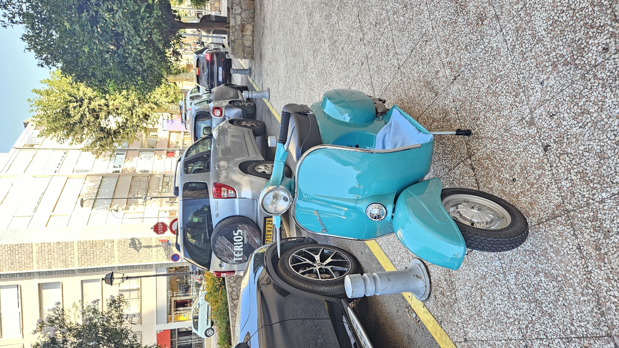 A turquoise Vespa scooter is parked on a textured concrete sidewalk next to a yellow line in L'Île-Rousse, France. The scooter has a black seat and a light blue towel draped over the front. Behind the scooter, a black SUV and several silver cars are parked along the street. The background shows a building with several floors, trees, and a stop sign. The sun is casting shadows, indicating it is likely late afternoon or early morning.