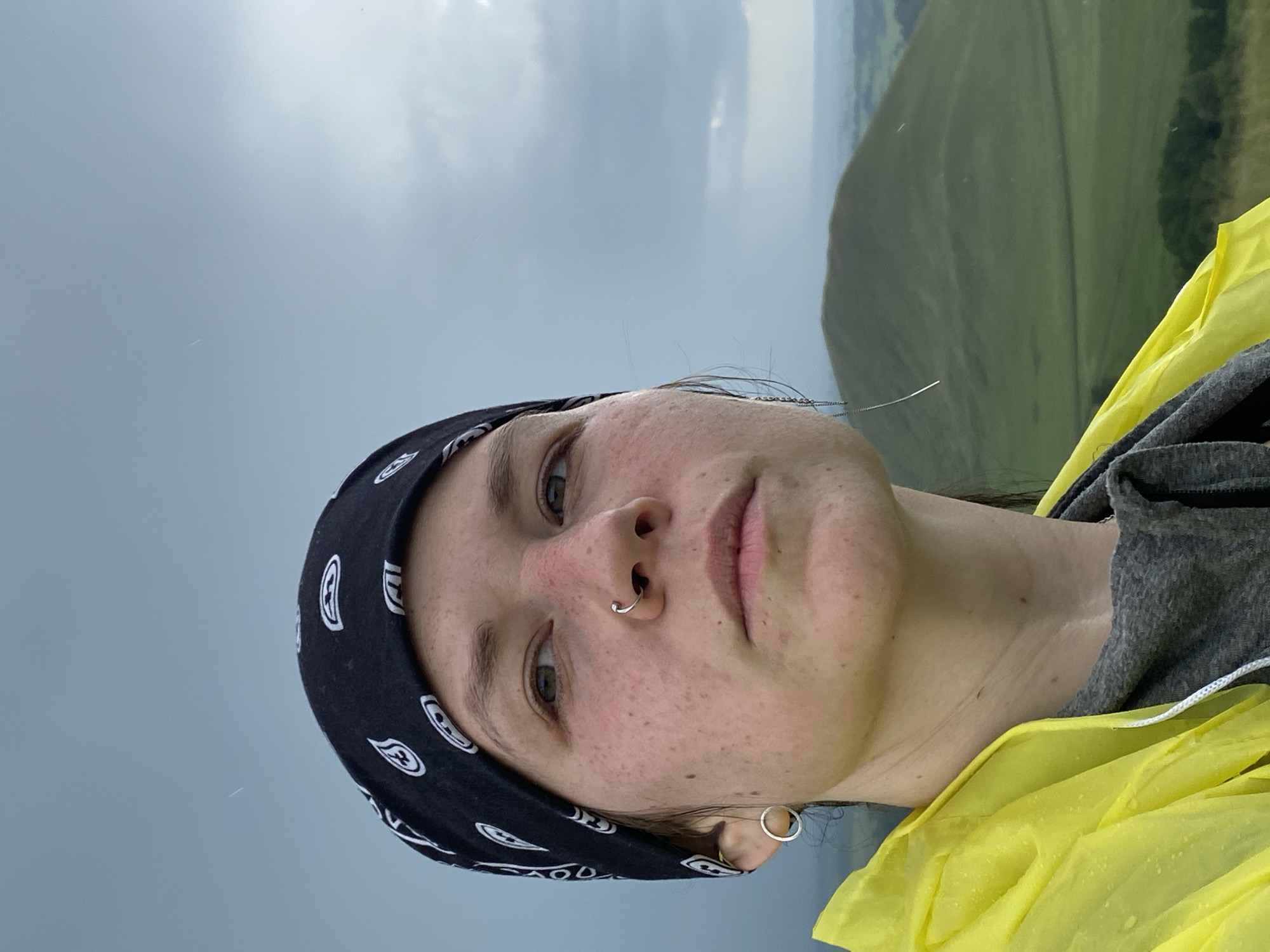 A close-up, slightly angled selfie shows a person wearing a black bandana with a white paisley pattern. They have a nose ring and a hoop earring in their left ear. The person is looking off to the right, with a neutral expression. They are wearing a bright yellow hooded jacket over a grey t-shirt. The background is a blurred landscape with rolling green hills under a stormy, overcast sky. Tiny white specks, possibly raindrops or insects, are visible in the air. The location is described as Рыскулово, Russia.