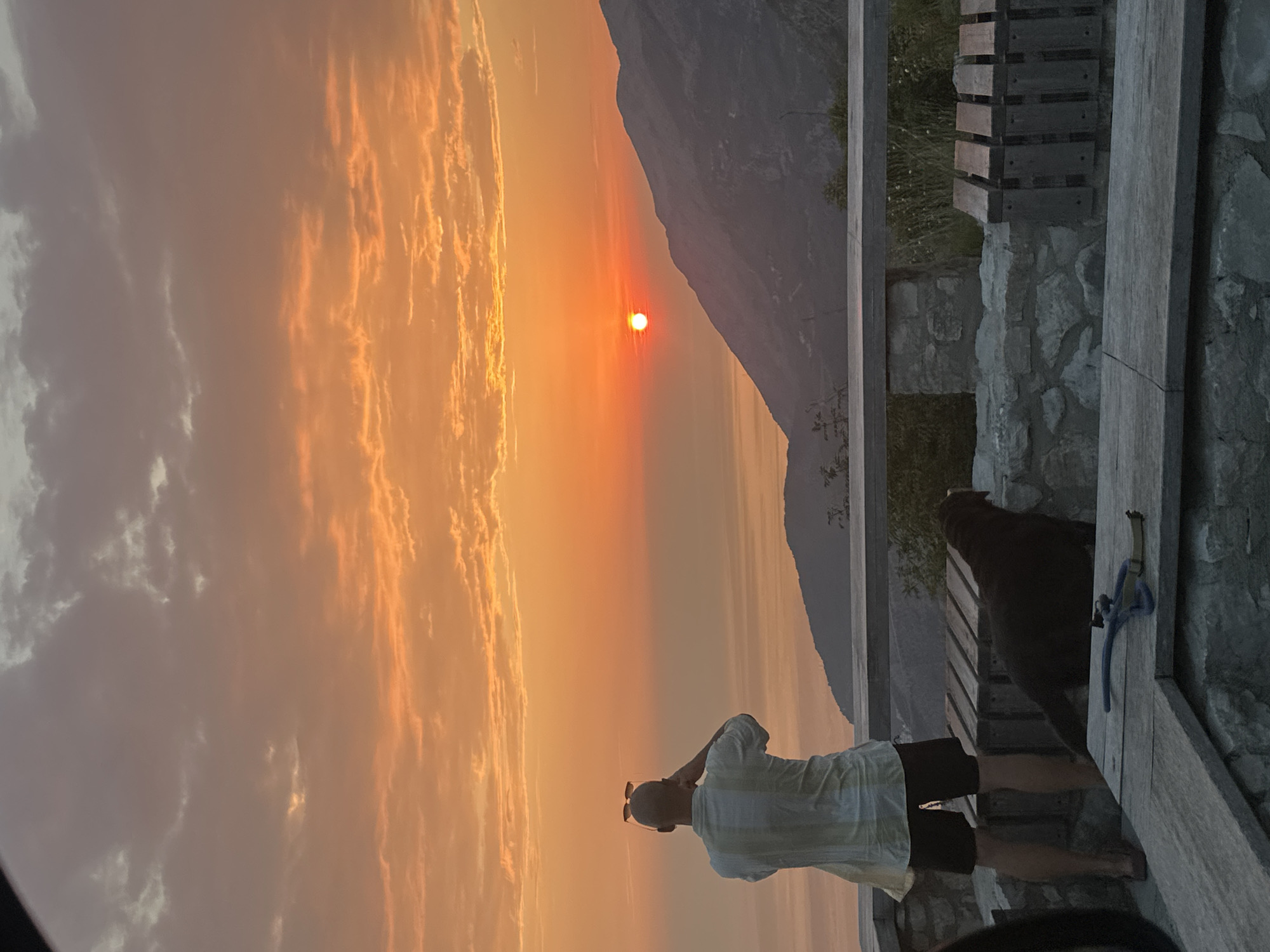At sunset in Gluhi Do, Montenegro, a man stands with his back to the camera, looking out at the ocean and mountains. He wears a striped shirt and dark shorts, with sunglasses perched on his head. A dark-colored dog sits beside him, also facing away from the viewer. They are positioned on a stone patio with wooden benches and railings, overlooking a body of water and a range of mountains. The sky is filled with orange and pink clouds, indicating the sun is setting. The scene conveys a sense of tranquility and natural beauty.