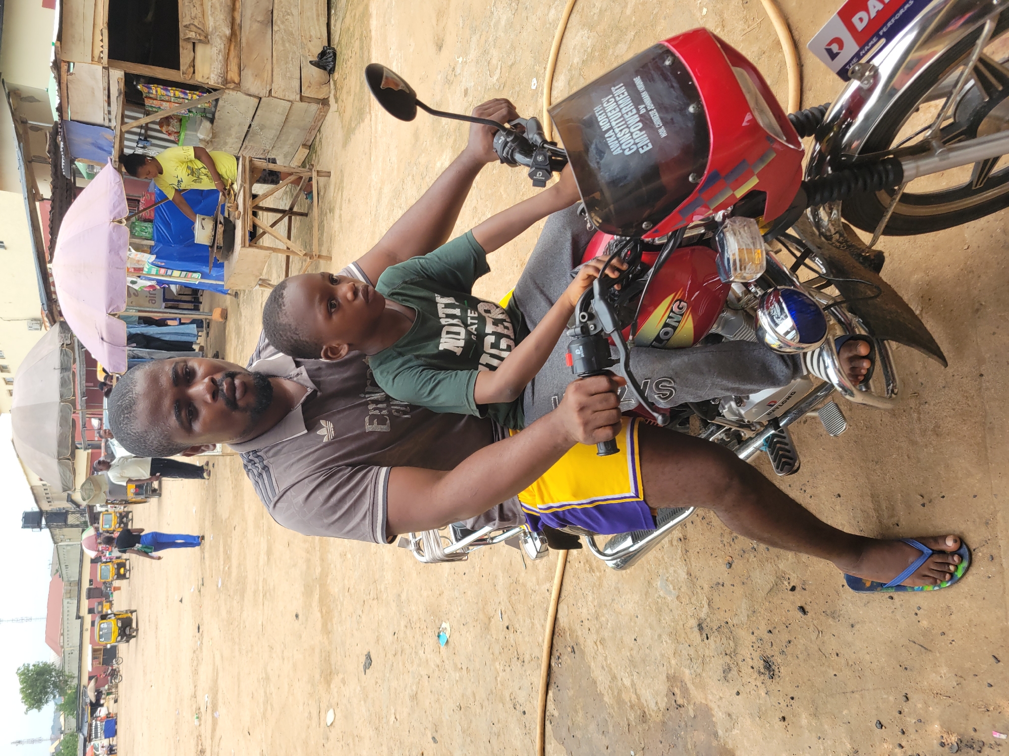 In Egbagu, Nigeria, a man and a young boy are seated on a red motorcycle in a dusty outdoor marketplace. The man, wearing a grey t-shirt and yellow and purple shorts, is in the driver's position, with his hands on the handlebars. The boy, in a green t-shirt, sits in front of him. The motorcycle has a red fairing with text that reads "AWKA NORTH CONSTITUENCY EMPOWERMENT" and "HON. CHIEZEUS JOHNMARK NWUNNE". In the background, several yellow tricycles, market stalls with umbrellas, and other people are visible. The ground is a mixture of dirt and debris.